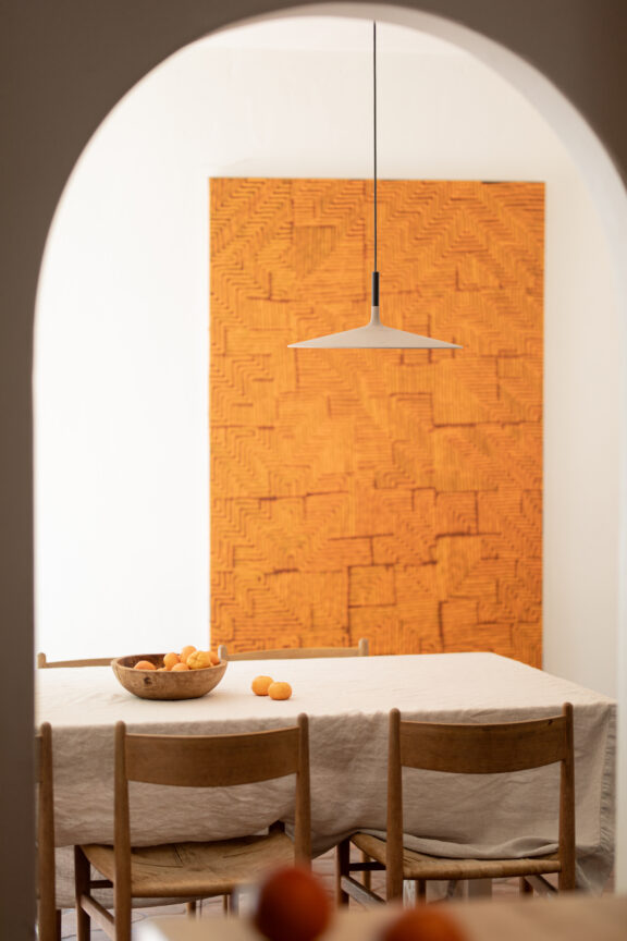 A dining table with a beige tablecloth, wooden chairs, a bowl of oranges, and a large orange textured artwork on the wall, viewed through an arched doorway.