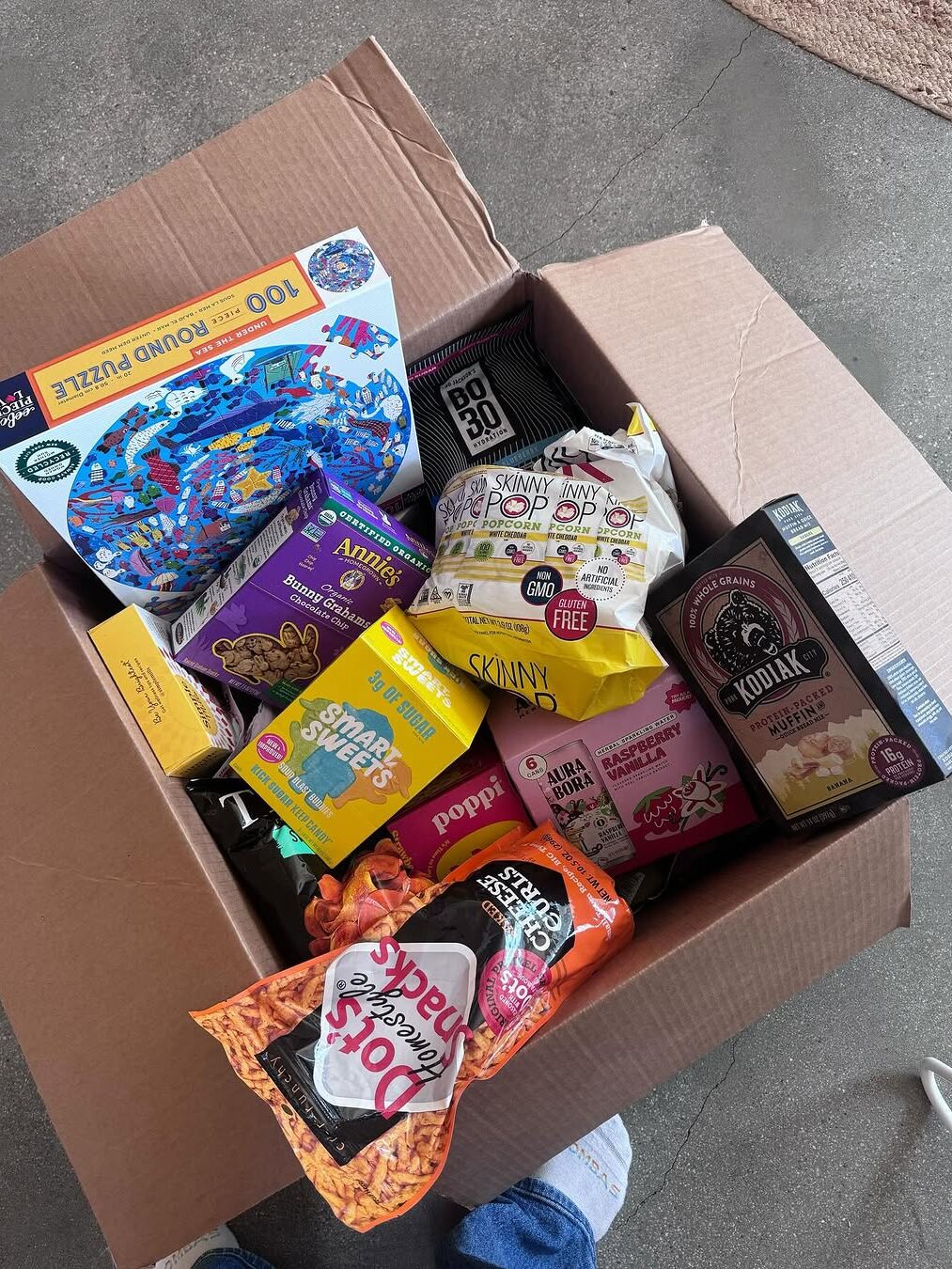 Open cardboard box filled with various snacks, a puzzle box, and a jigsaw puzzle.