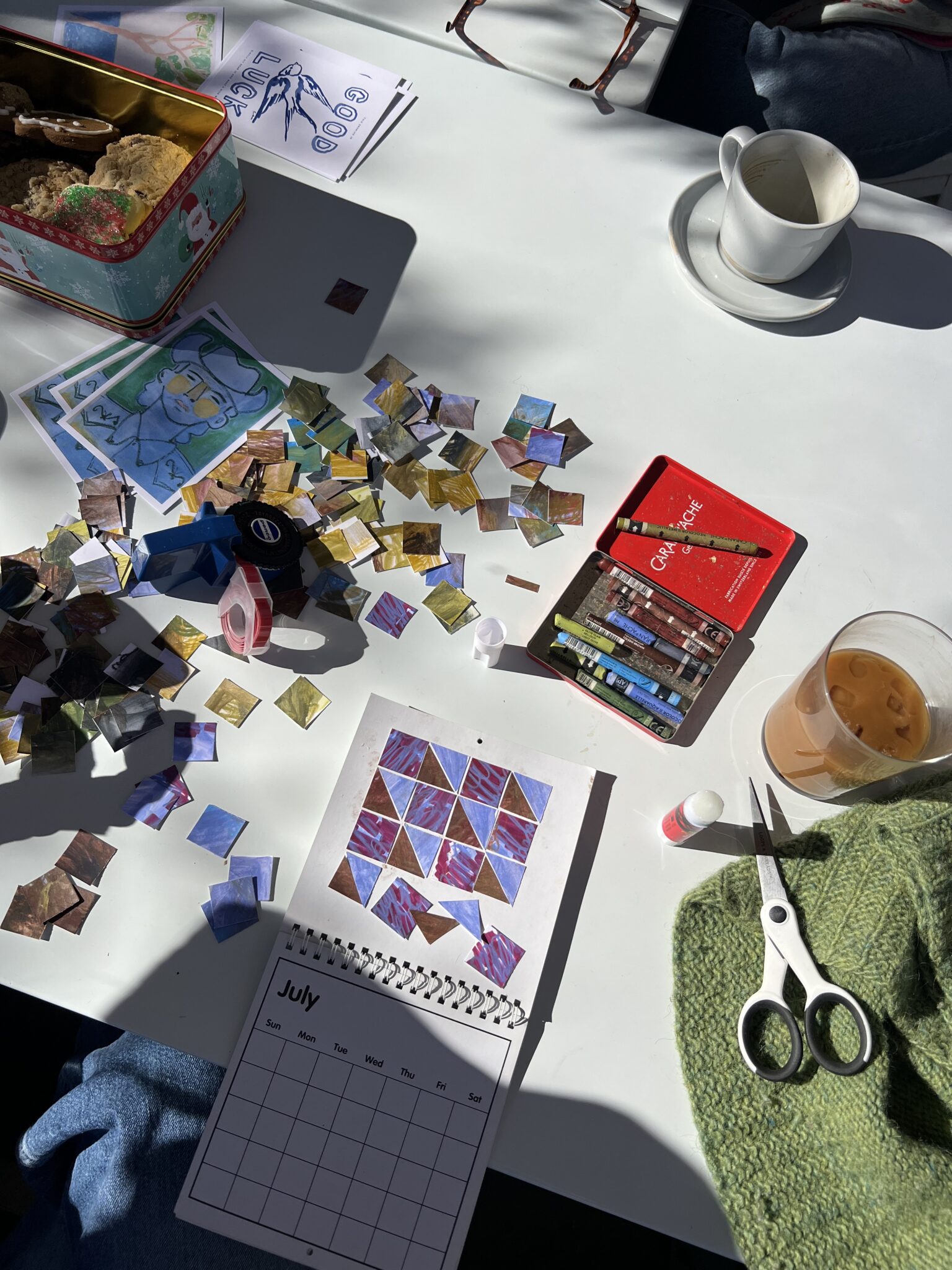 A table with colored paper squares, oil pastels, scissors, glue, a July calendar, a cup, a drink, and snacks, with people’s hands partially visible around the table.