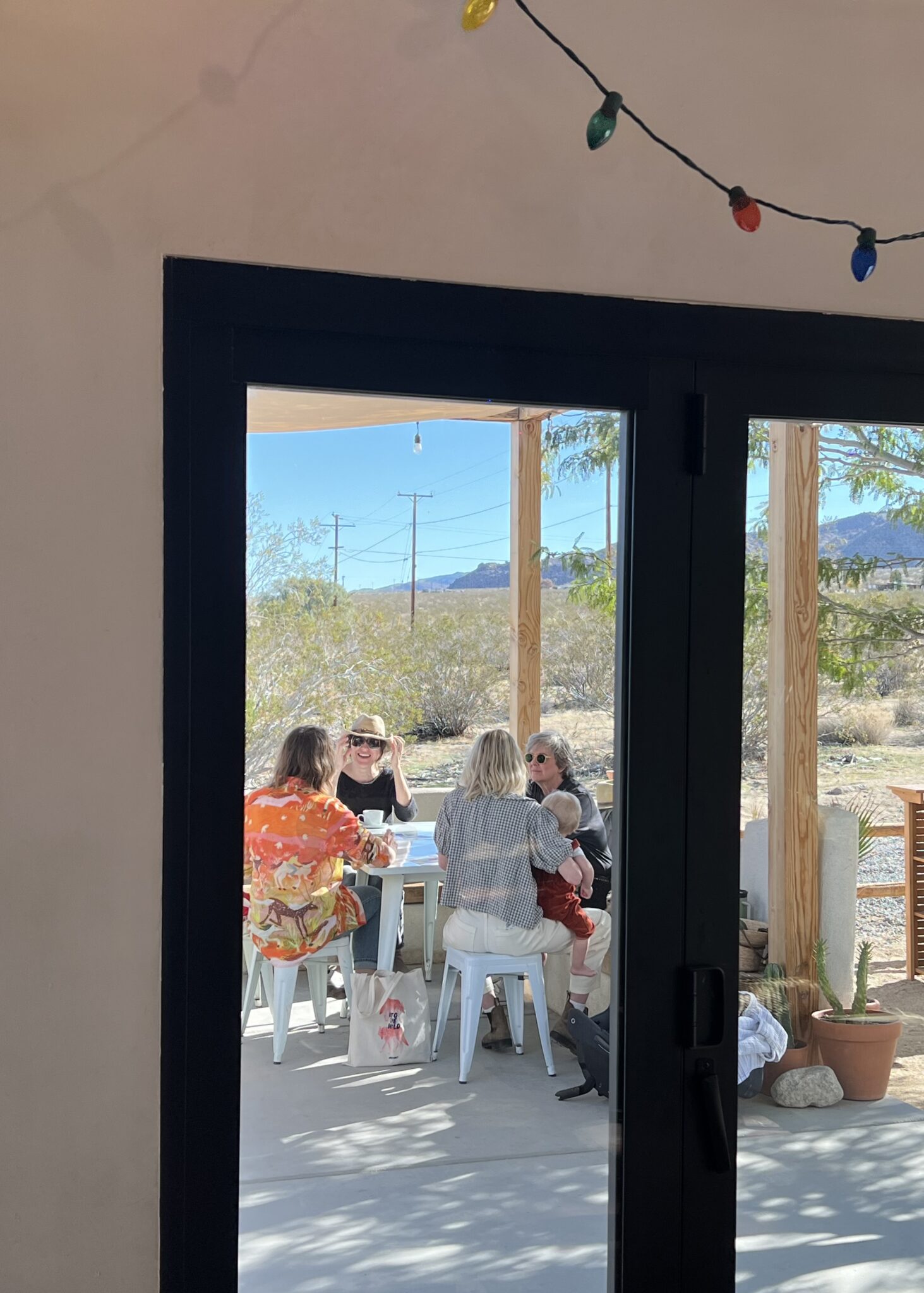 Four adults and a child sit around a patio table outdoors, seen through a glass door, with desert landscape and string lights visible.