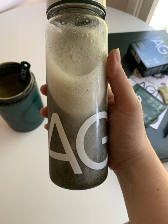 A hand holds a clear bottle filled with a frothy green liquid. The letters "AG" are visible on the bottle. A container and packets are on the white table.