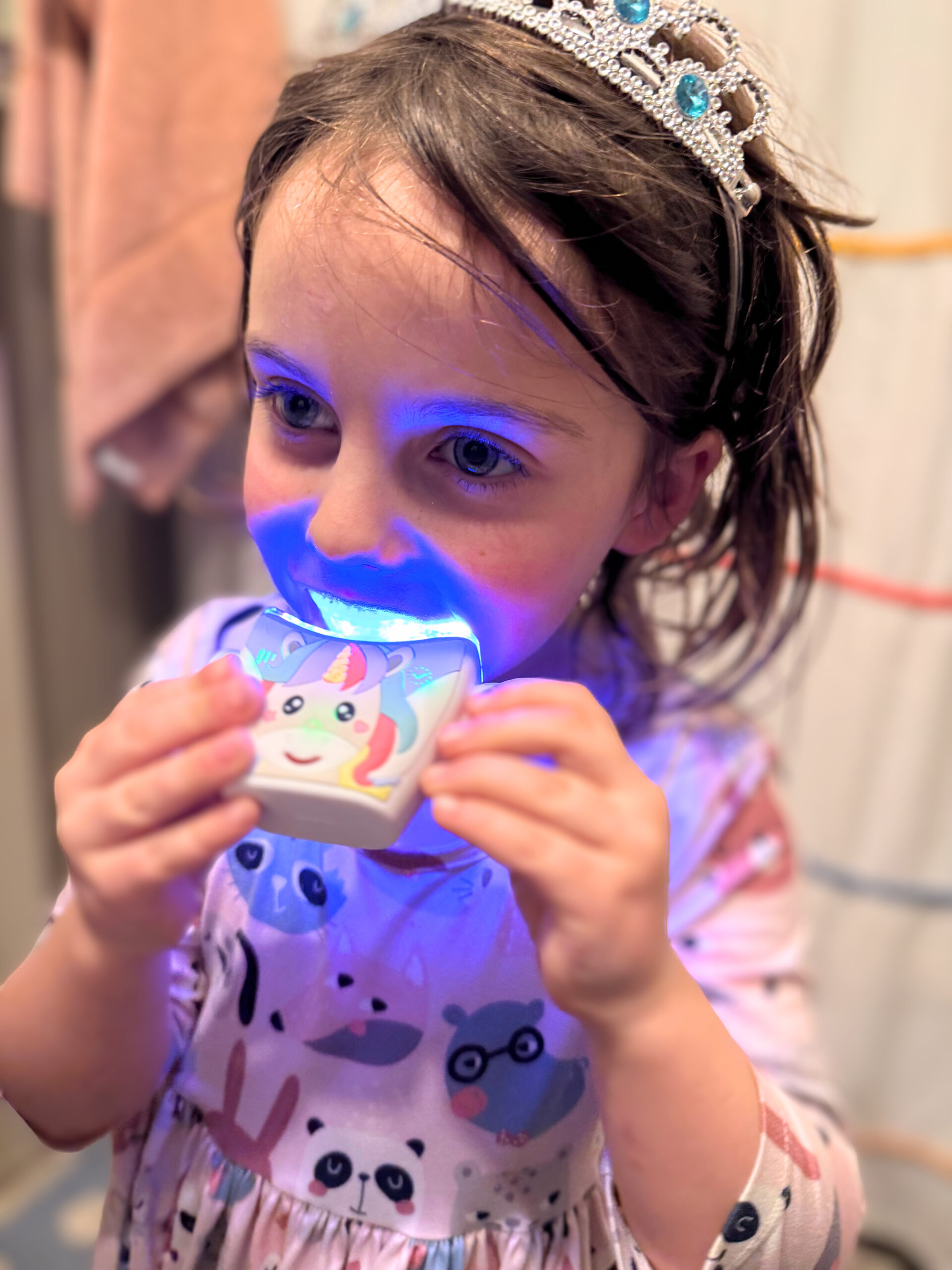 Child wearing a tiara uses a lighted dental device with a unicorn design, wearing pink pajamas with animal prints.