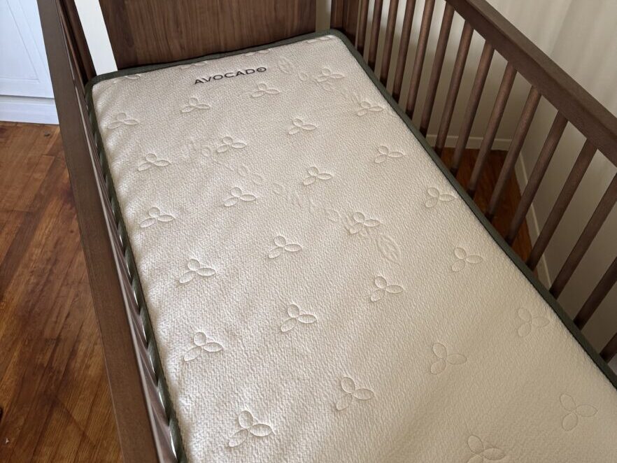 A wooden baby crib with a white Avocado brand organic mattress inside, positioned on a hardwood floor next to a cream-colored wall.