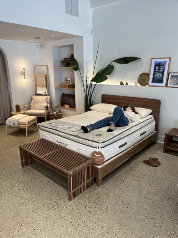 A person lies on a mattress in a modern showroom with wooden furniture, a large plant, framed art on the wall, and a lounge chair in the corner.