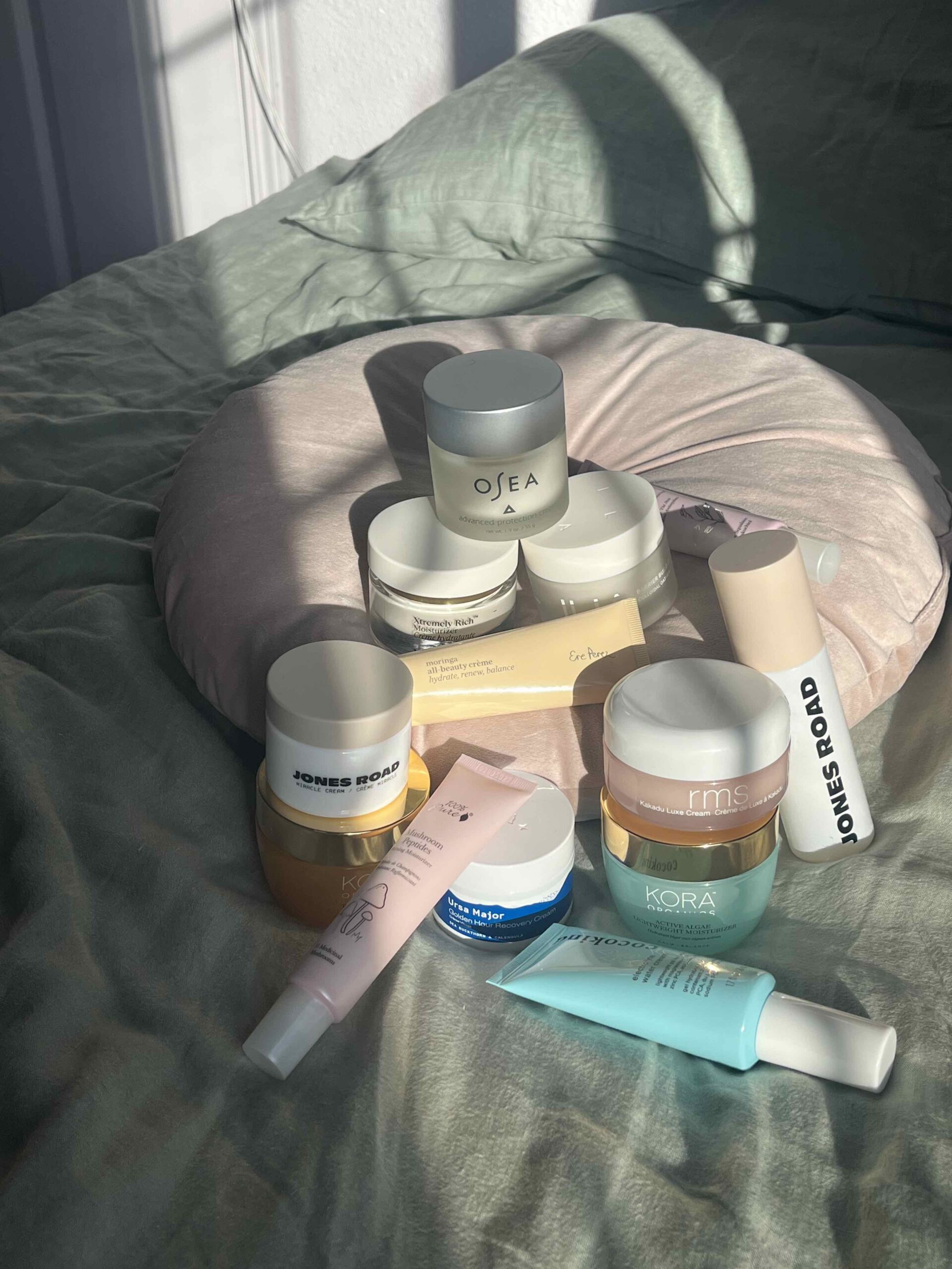 A collection of skincare products, including jars and tubes from brands like OSEA, Jones Road, and Kora, arranged on a bed with a round pink pillow in sunlight.
