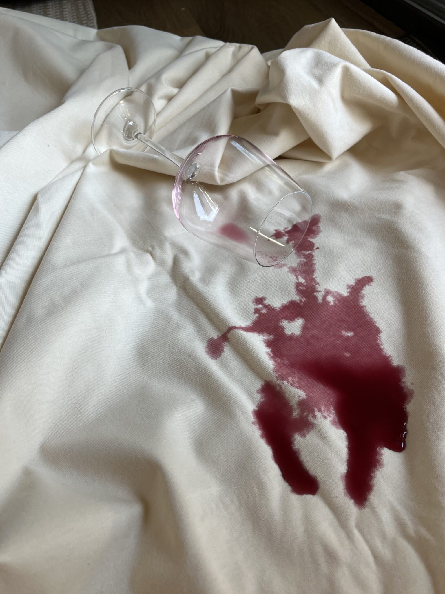 A wine glass lies on its side on a white cloth, with red wine spilled around it and staining the fabric.