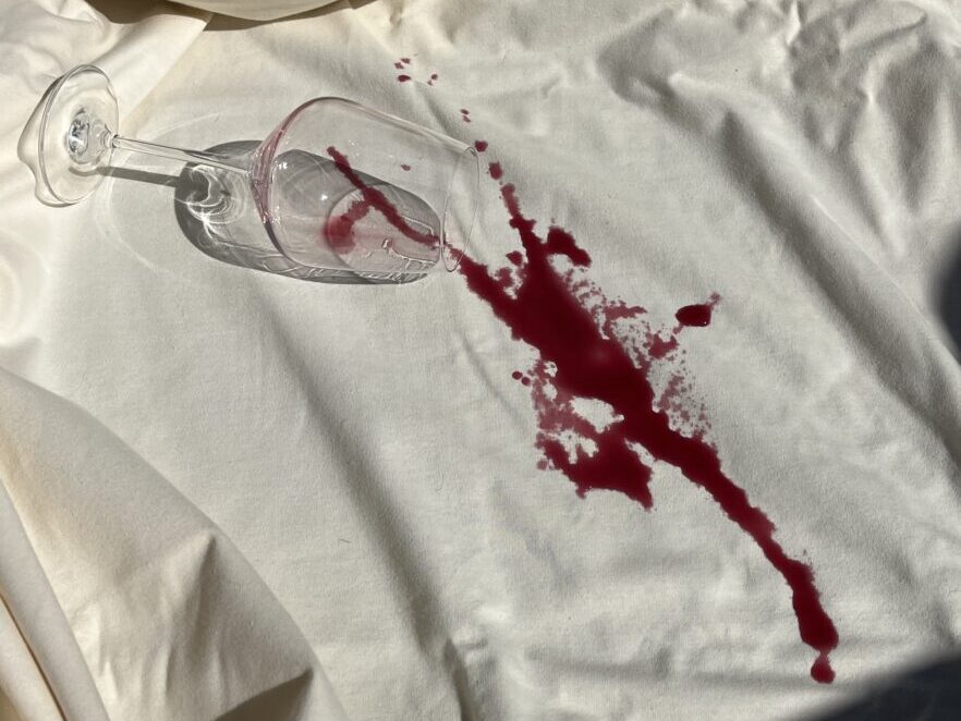 An overturned wine glass with red wine spilled across a white, wrinkled organic mattress surface.