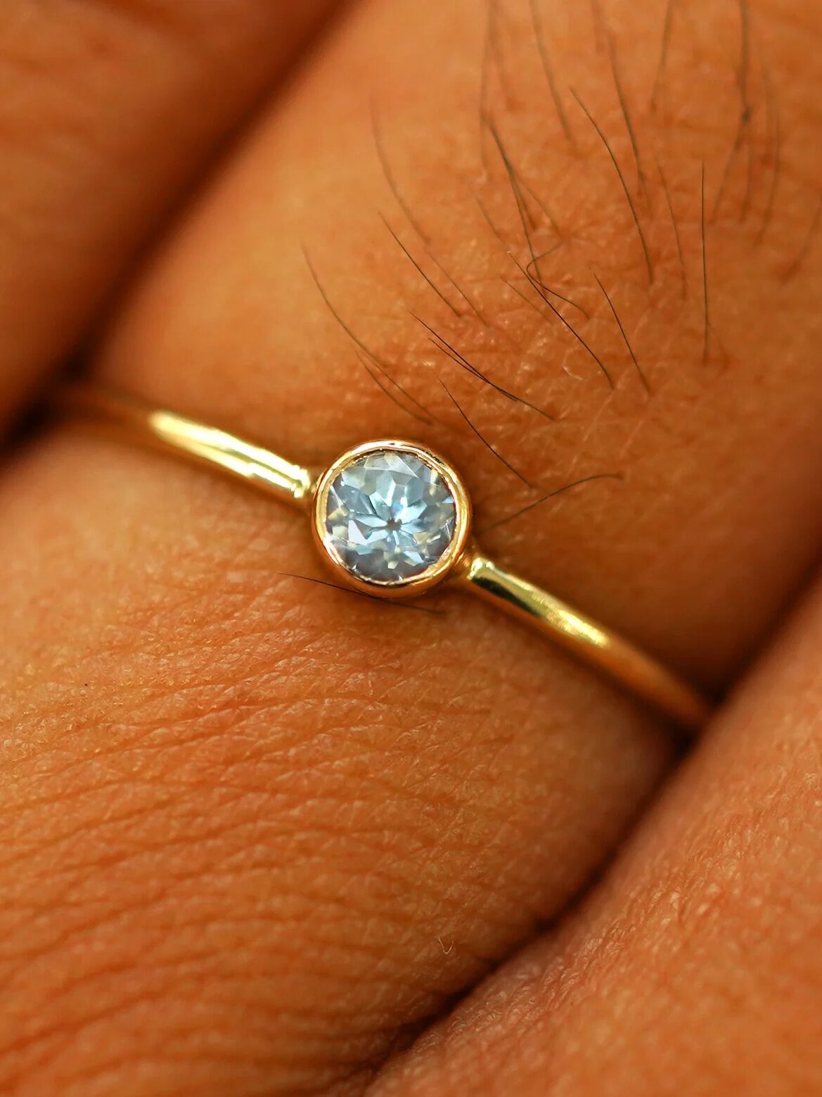 A close-up of a gold ring with a round, light blue gemstone set in the center, worn on a finger.
