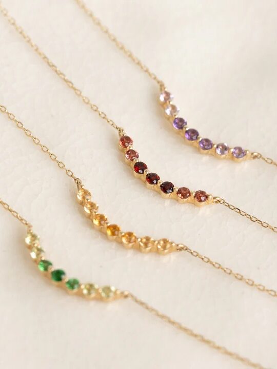 Four delicate gold chain necklaces, each with a curved row of small, colored gemstones in green, yellow, red, and purple, displayed on a light textured surface.
