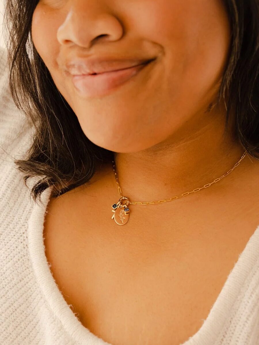A woman wearing a white sweater smiles slightly, showing a gold chain necklace with a small oval pendant and a charm.