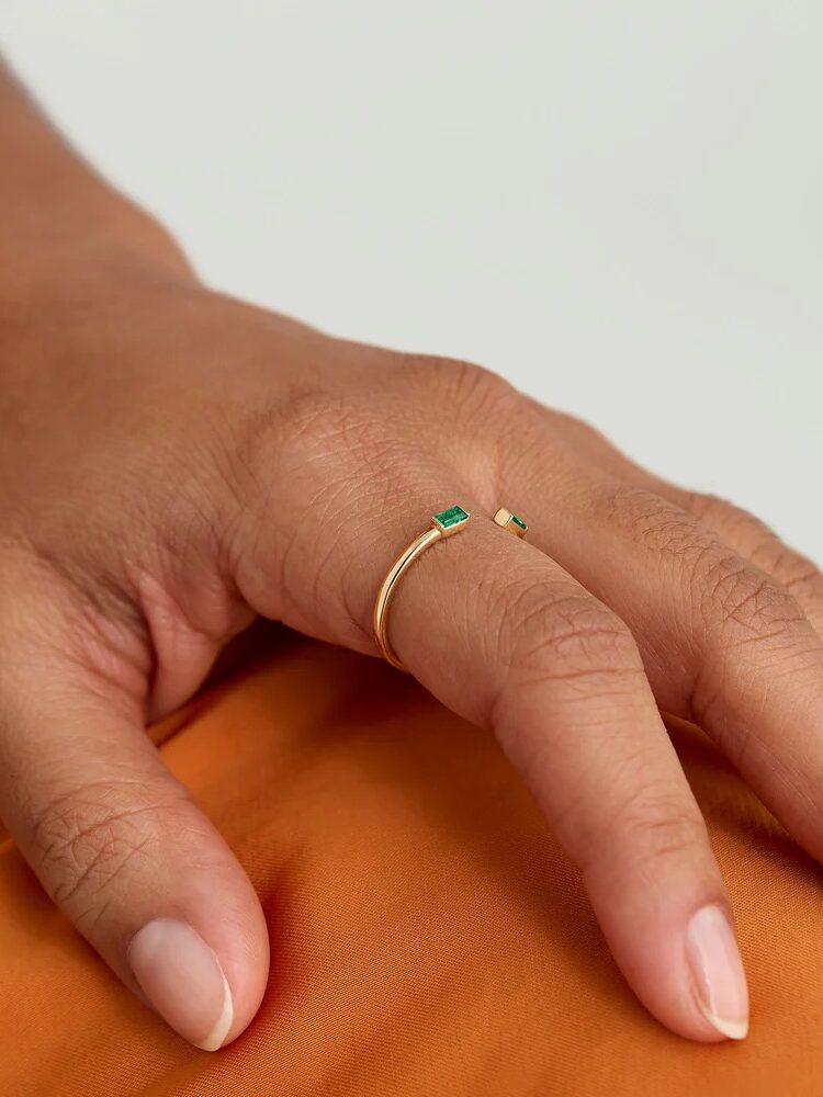 A hand with a gold open ring featuring green gemstones on the pinky finger, resting on an orange fabric.