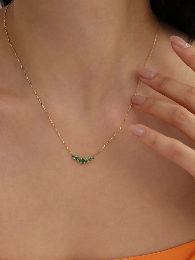 A close-up of a person wearing a delicate gold chain necklace with small green stones, touching their collarbone with manicured fingers.