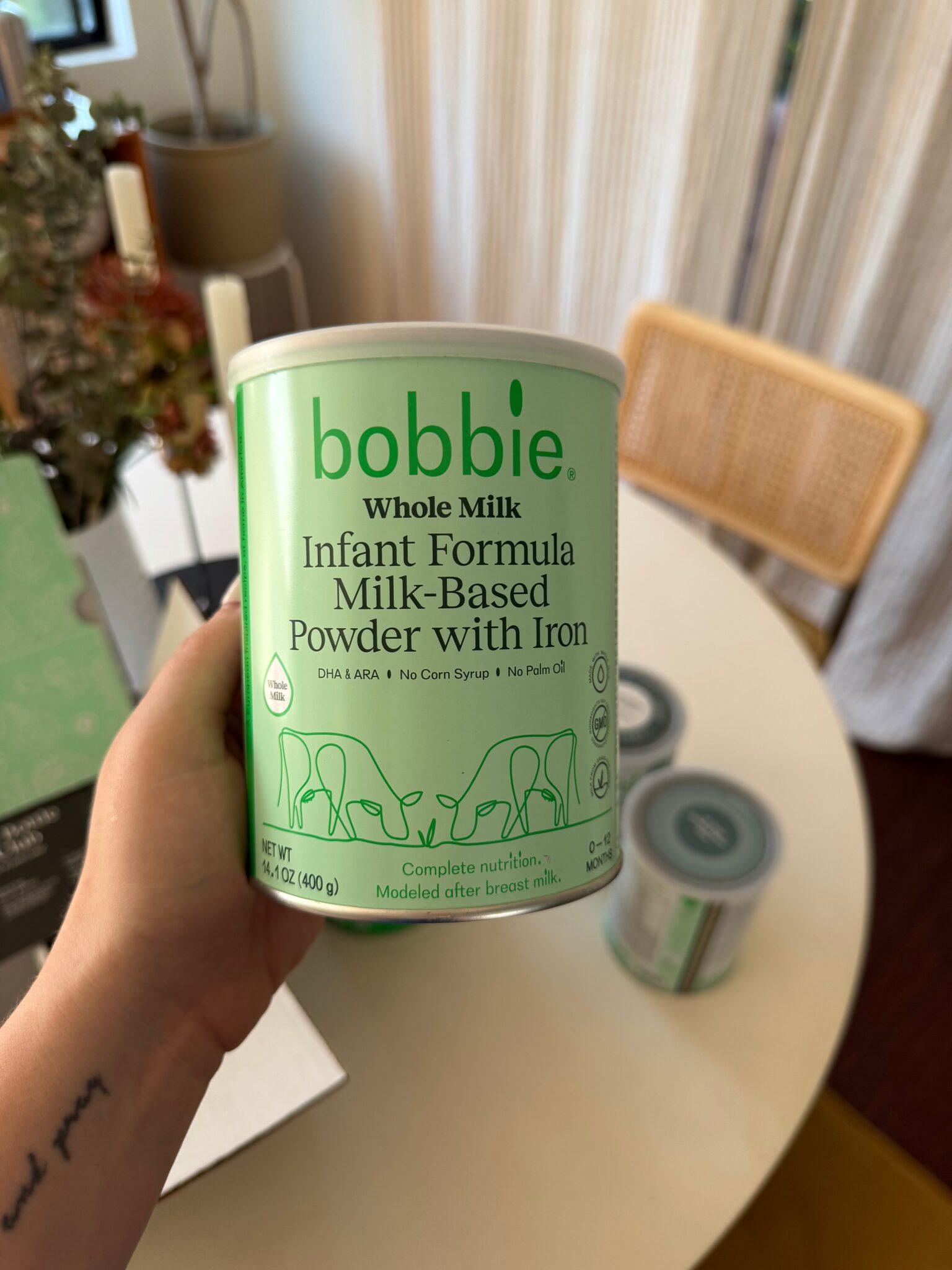 A hand holds a can of Bobbie Whole Milk Infant Formula, with two more cans on a round table in the background. The label highlights key features of the formula.