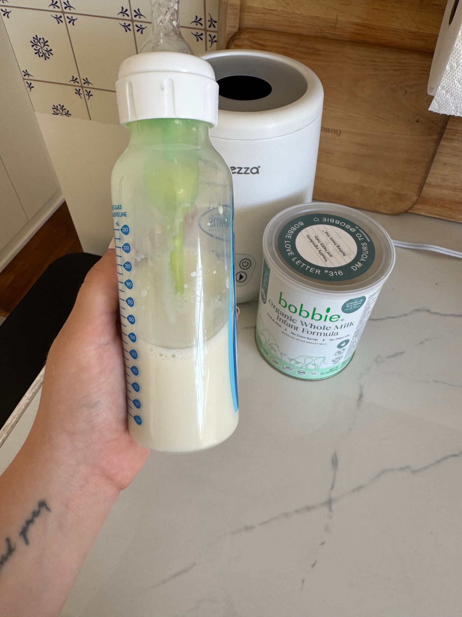 A hand holds a baby bottle filled with milk in front of an open can of Bobbie organic whole milk infant formula on a kitchen counter.