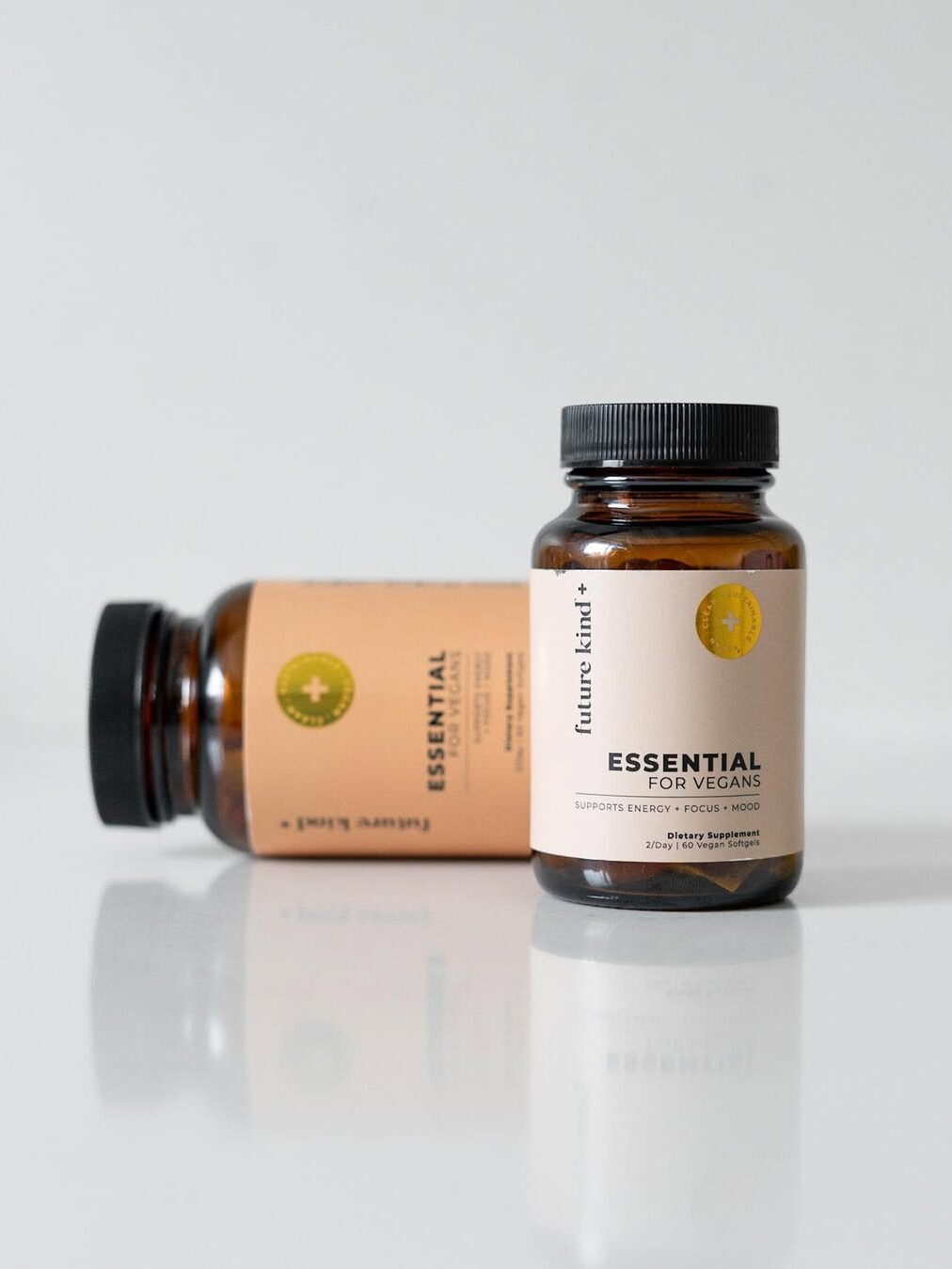 Two brown bottles with beige labels displaying "Essential for Vegans" on a white reflective surface. One bottle stands upright, and the other lies on its side.