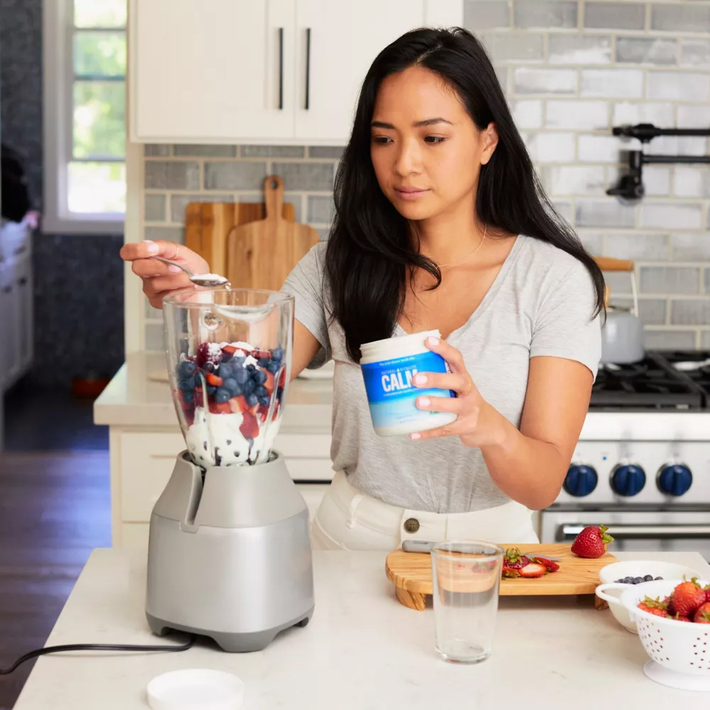 A woman drops a spoonful of Calm calcium supplement powder into a blender. 