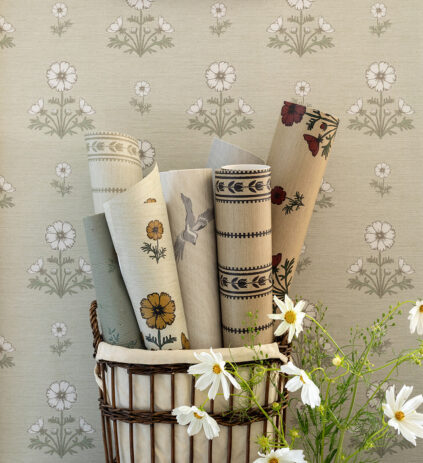 A basket holds rolled wallpaper with floral and bird patterns, set against a wall with daisy prints. White daisies are in the foreground.