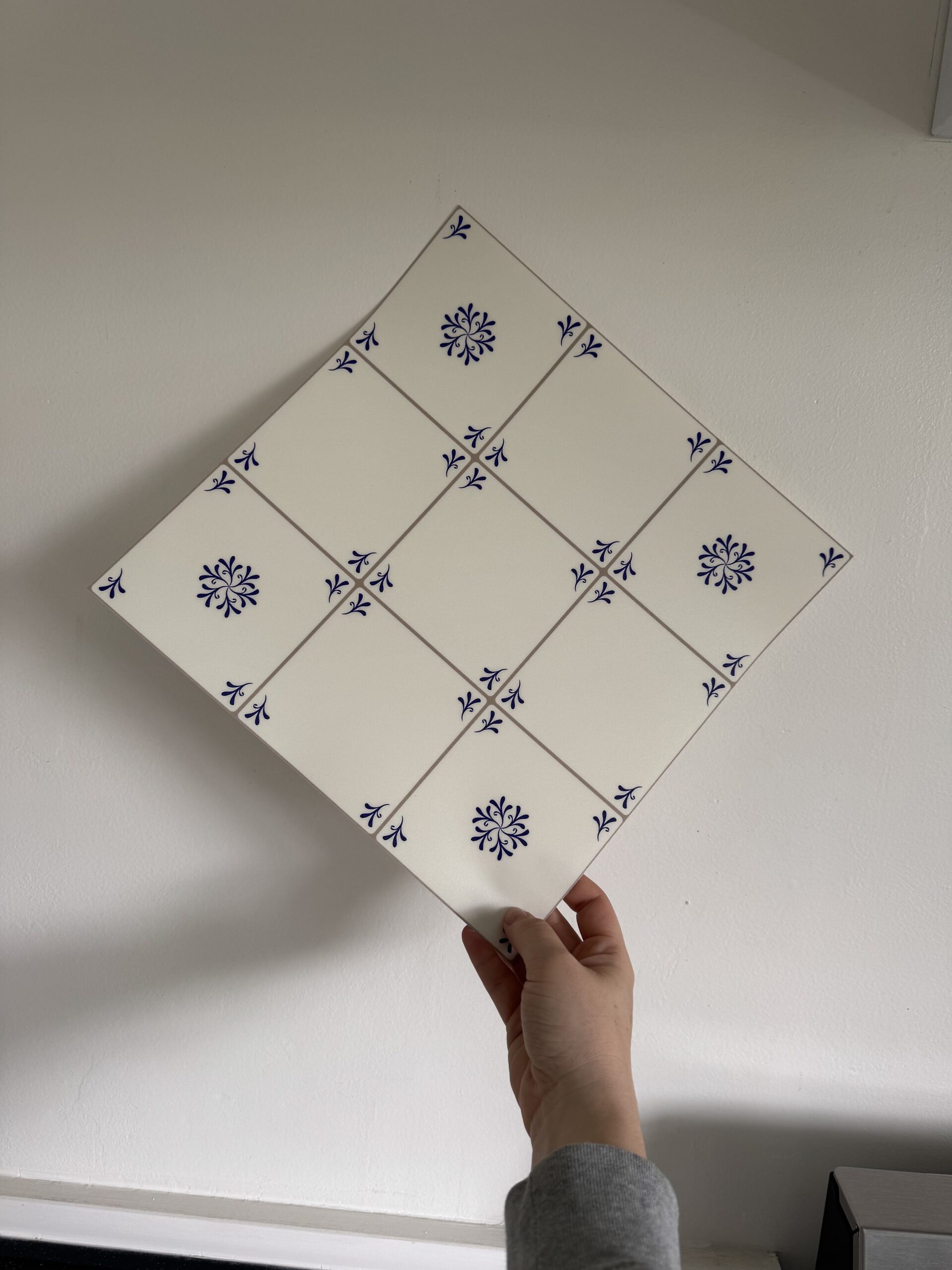 A hand holds a large ceramic tile composed of nine smaller white tiles with blue floral patterns, arranged in a 3x3 grid.