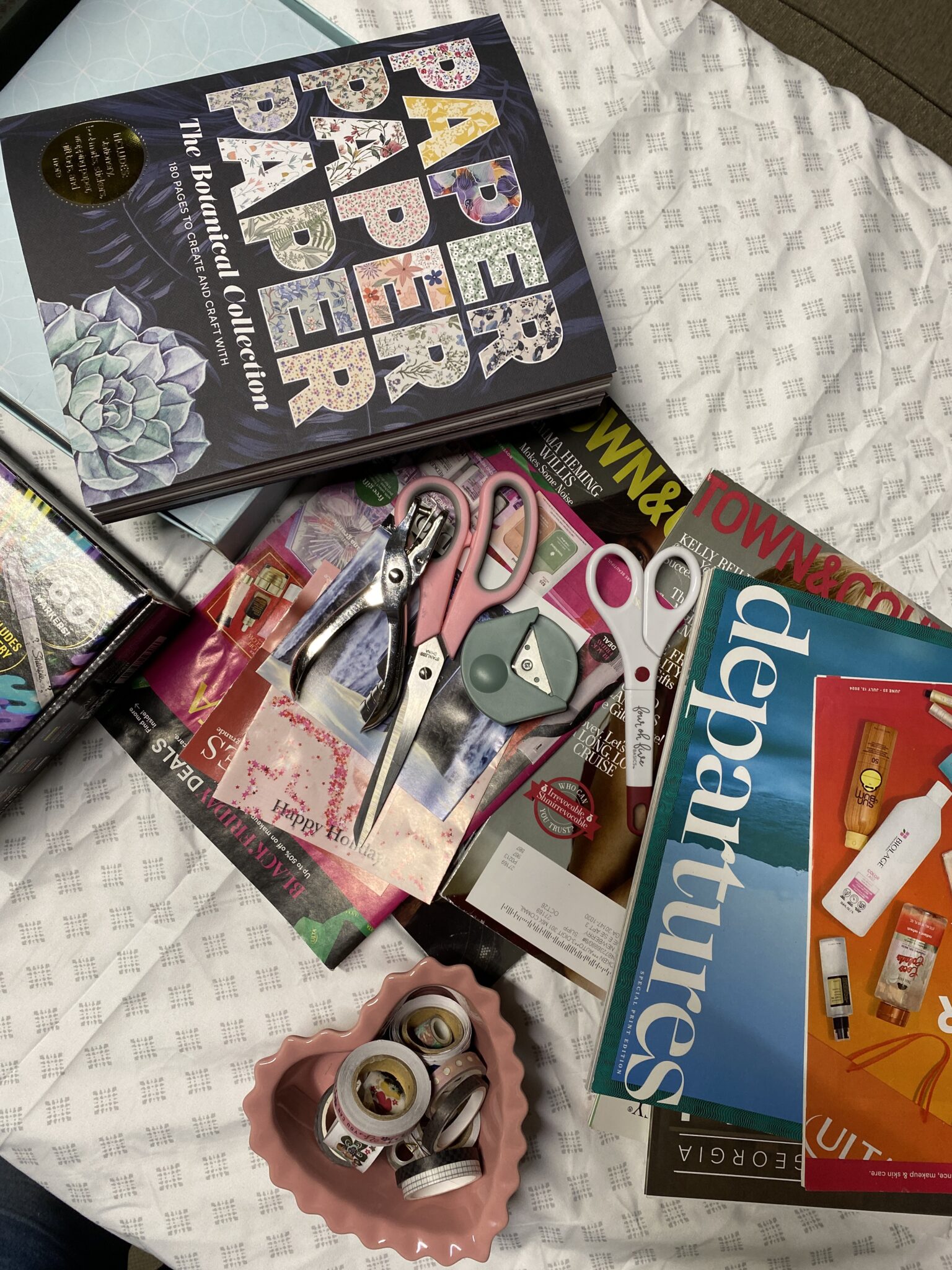 Assorted craft supplies, scissors, magazines, washi tape, and a book titled "Paper Paper Paper" are spread out on a patterned fabric surface.