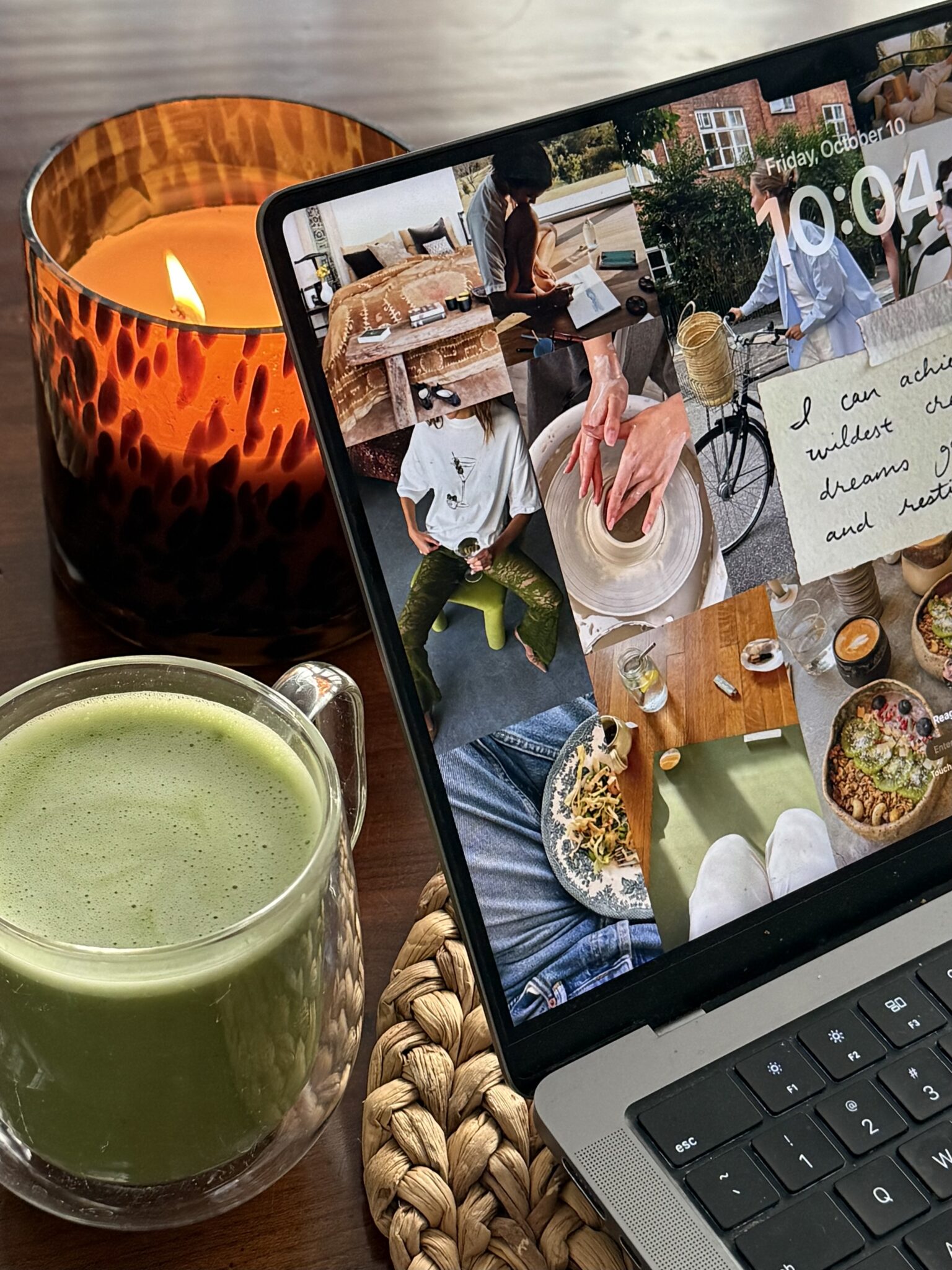 A glass mug of green matcha latte, a lit amber candle, and a laptop displaying a collage-style lock screen on a wooden table.