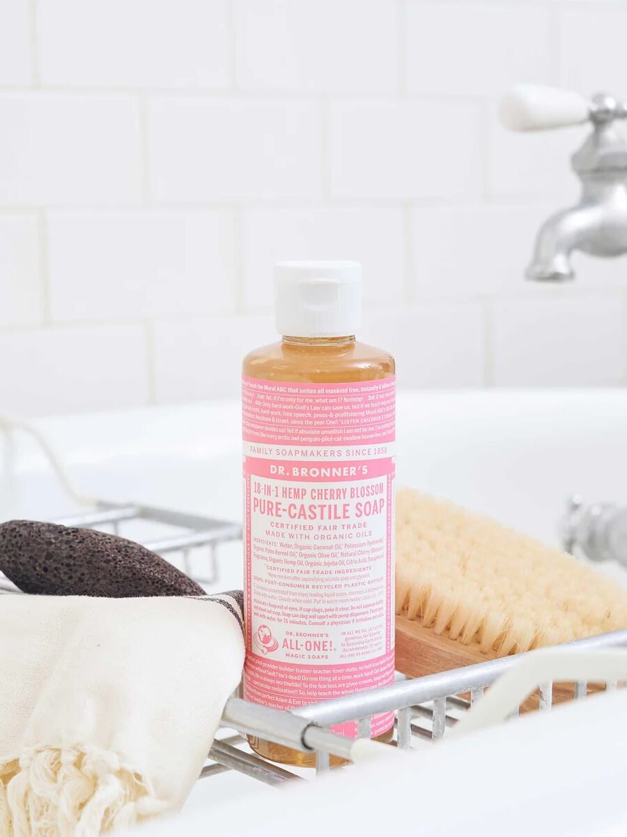 A bottle of Dr. Bronner’s 18-in-1 Hemp Cherry Blossom Pure-Castile Soap sits in a wire rack over a bathtub next to a pumice stone, a wooden bath brush, and a cloth.