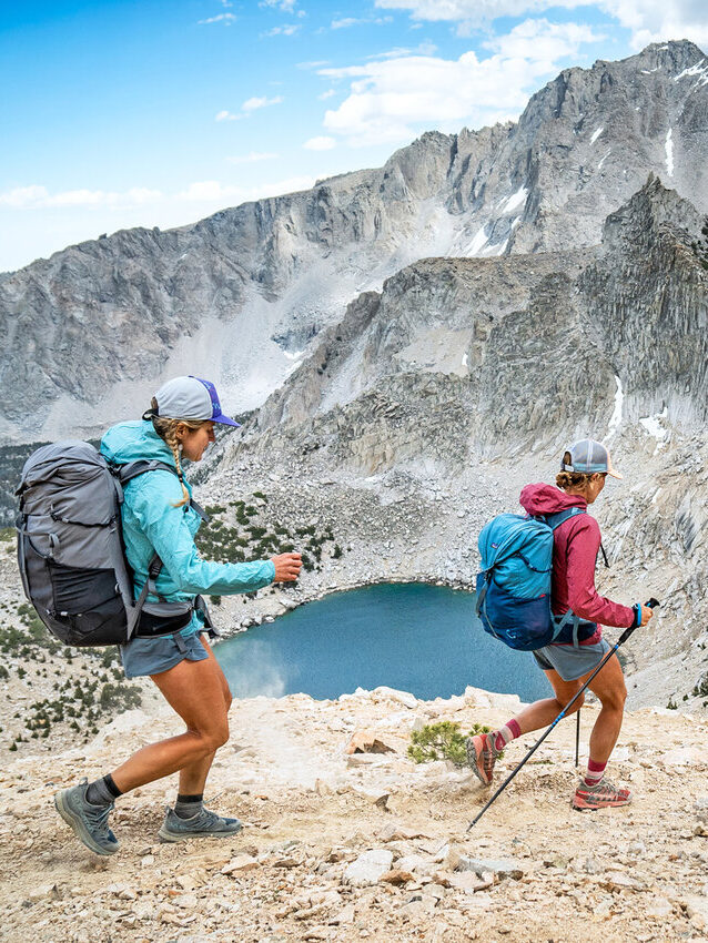 Two hikers with backpacks and trekking poles walk down a rocky mountain trail overlooking a blue alpine lake and rugged peaks under a partly cloudy sky.