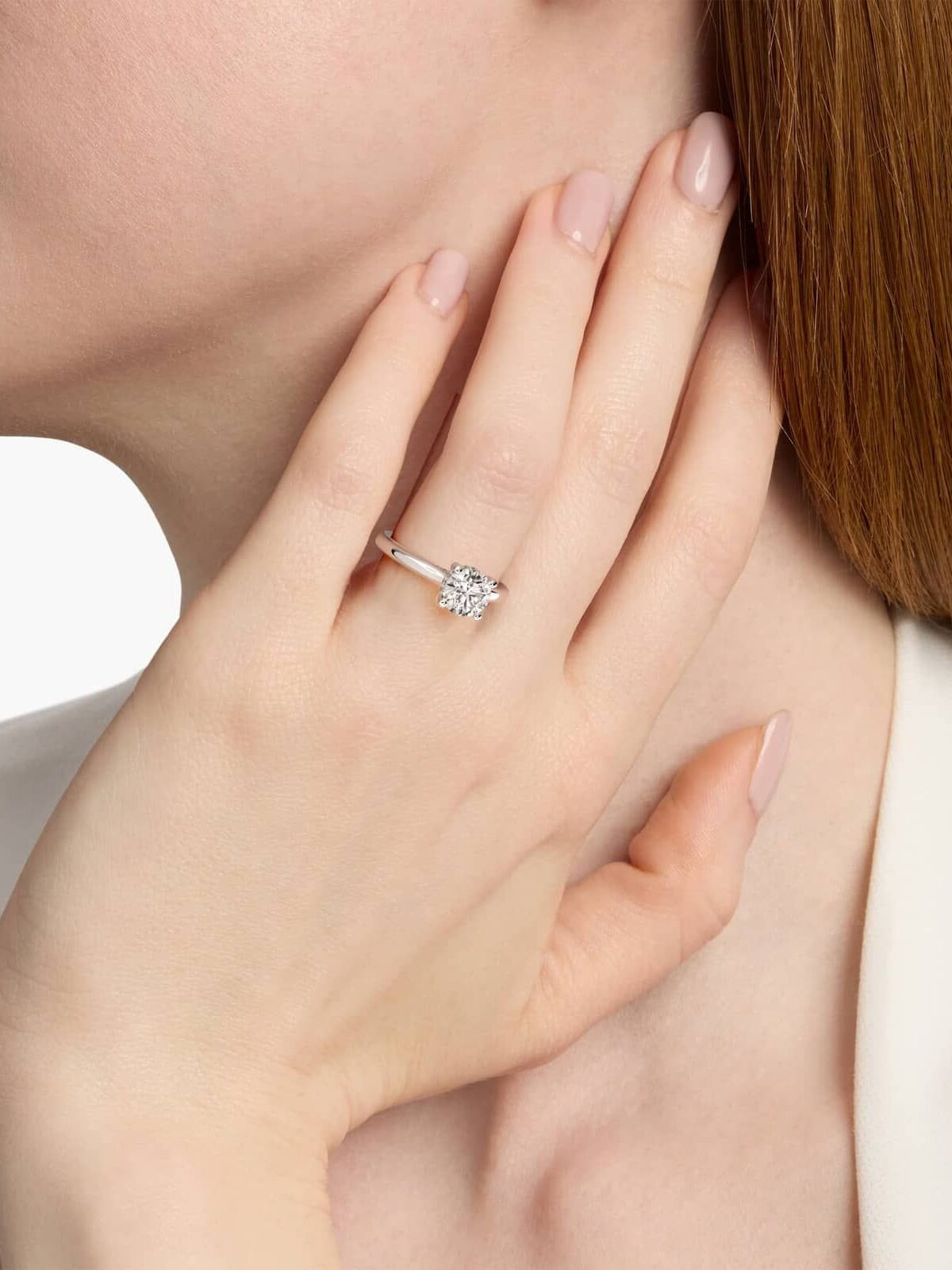 A woman with light skin wears a diamond solitaire ring on her left ring finger, touching her face with a relaxed hand.