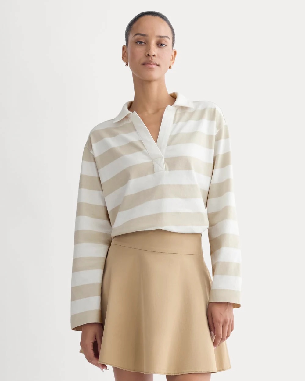 A woman wearing a beige and white striped long-sleeve shirt and a beige A-line skirt stands against a plain light background.