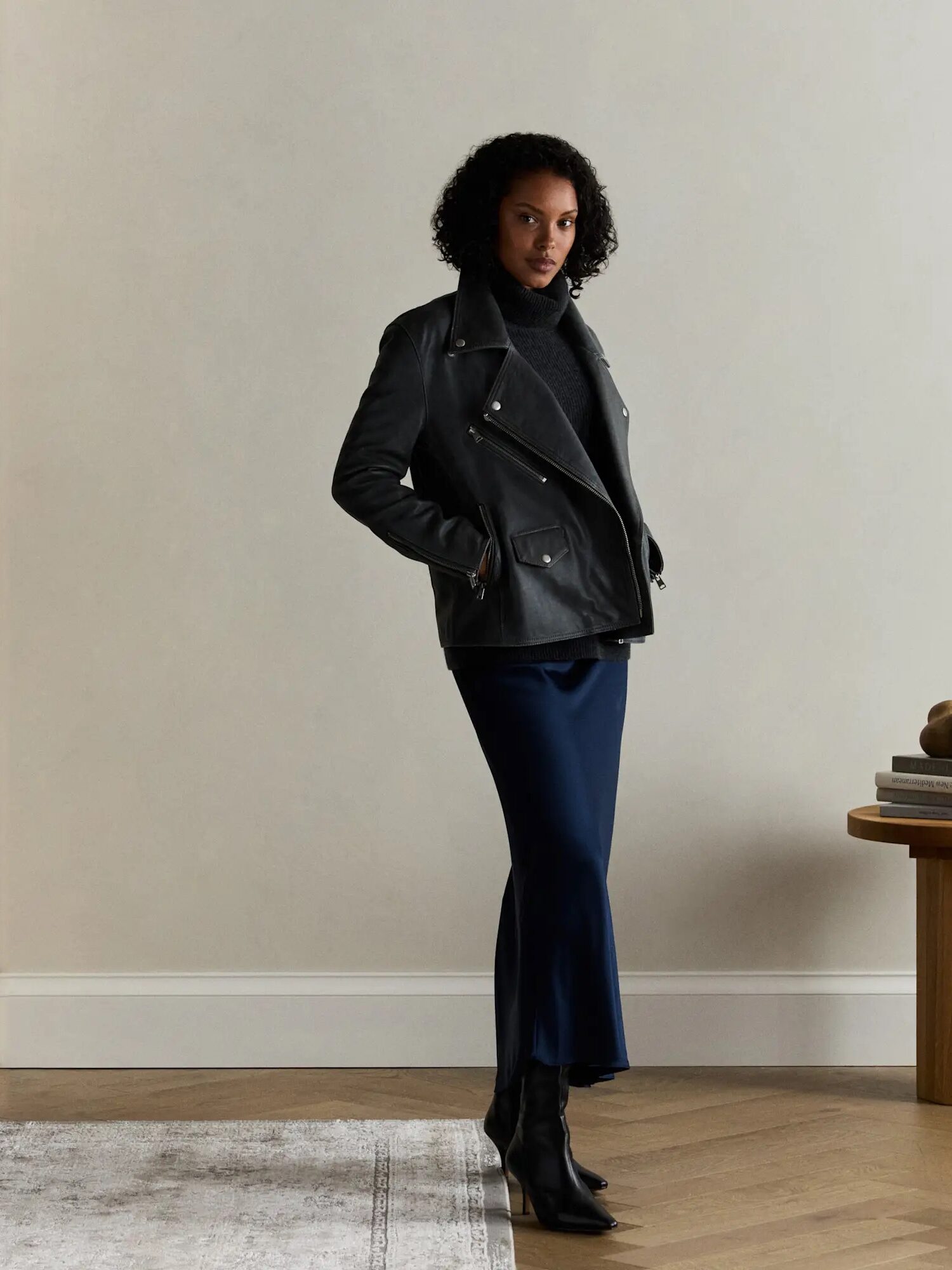 A woman stands indoors wearing a dark jacket, dark turtleneck, blue midi skirt, and black ankle boots, with her hands in her pockets.