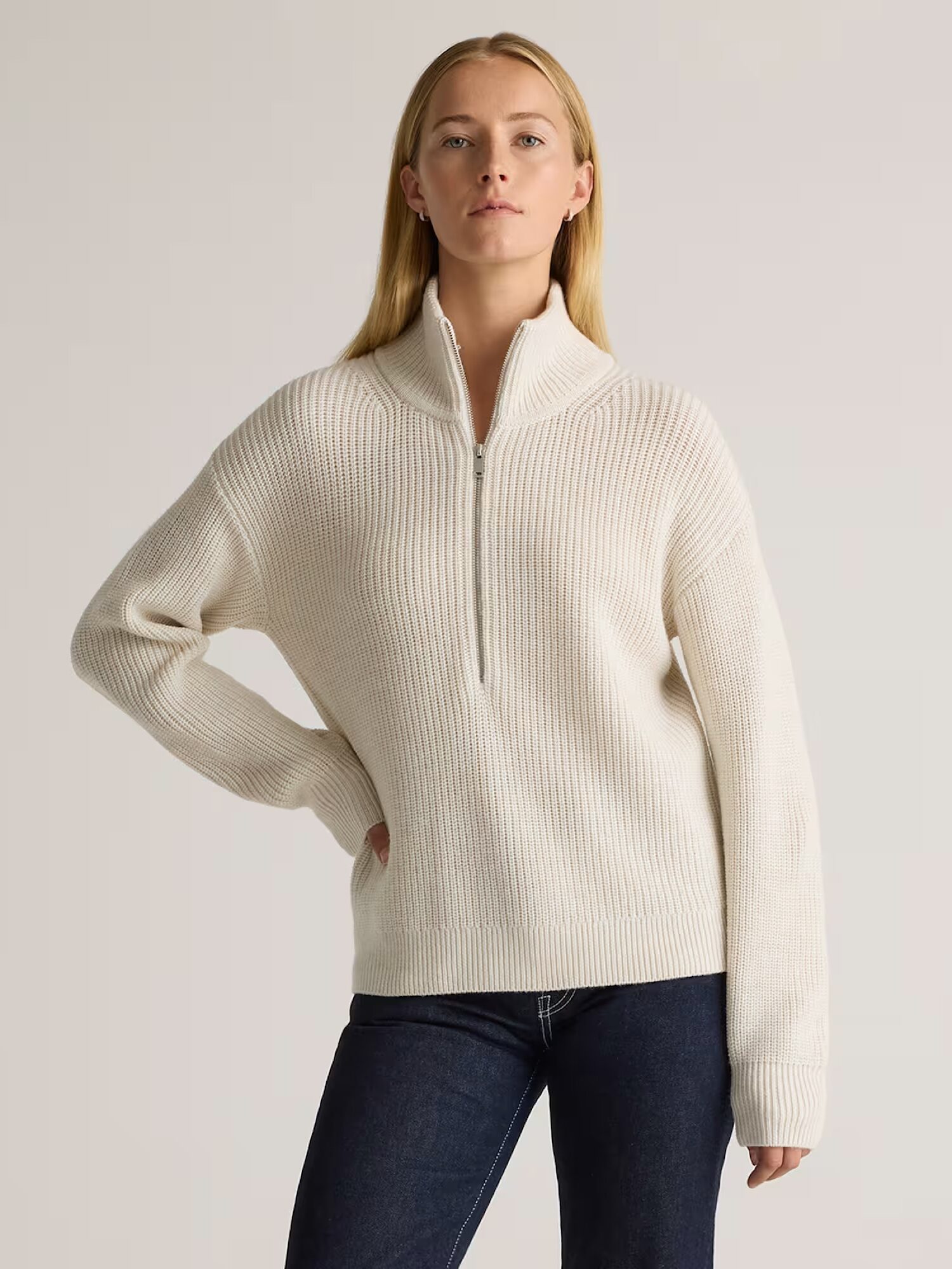 A person wearing a cream-colored, ribbed quarter-zip sweater and dark blue jeans stands against a plain light background.