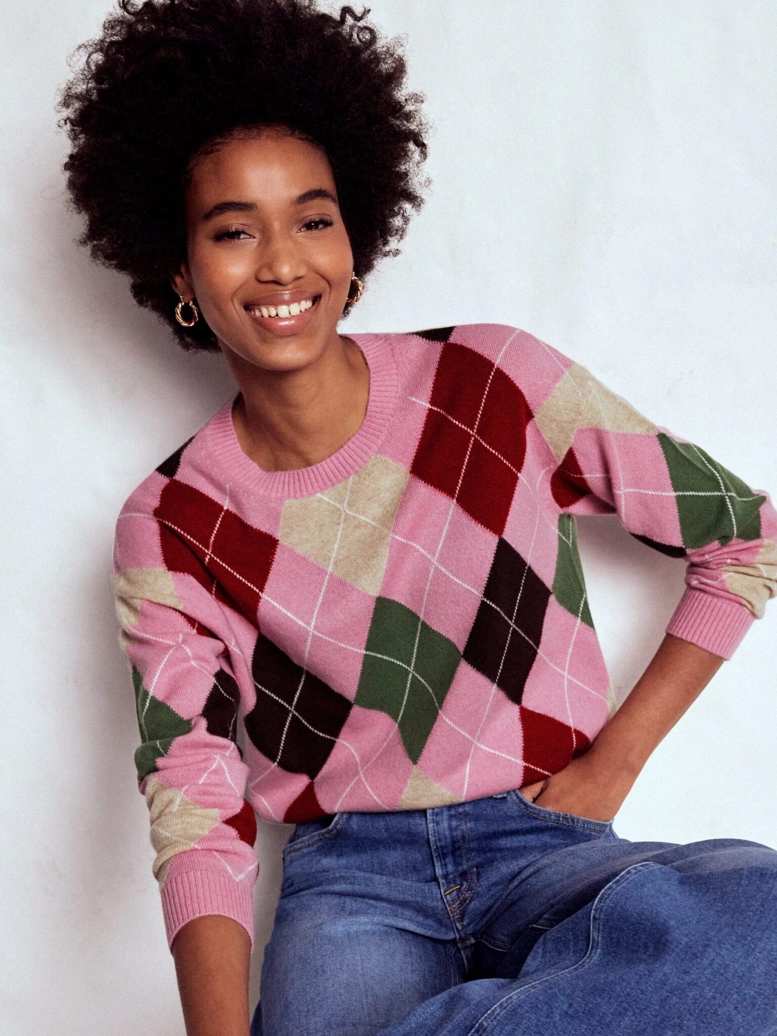 A person wearing a pink, green, red, and beige argyle sweater with blue jeans sits against a white background and smiles.