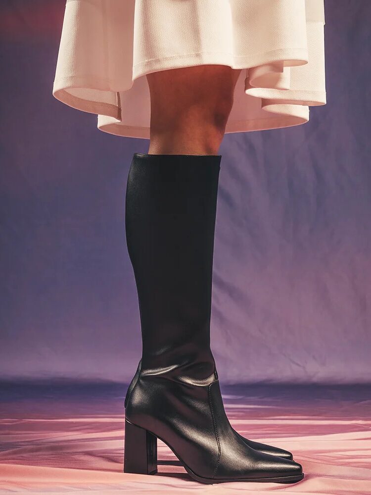 A person wearing a white skirt and black knee-high heeled boots stands on a pink surface with a purple backdrop.