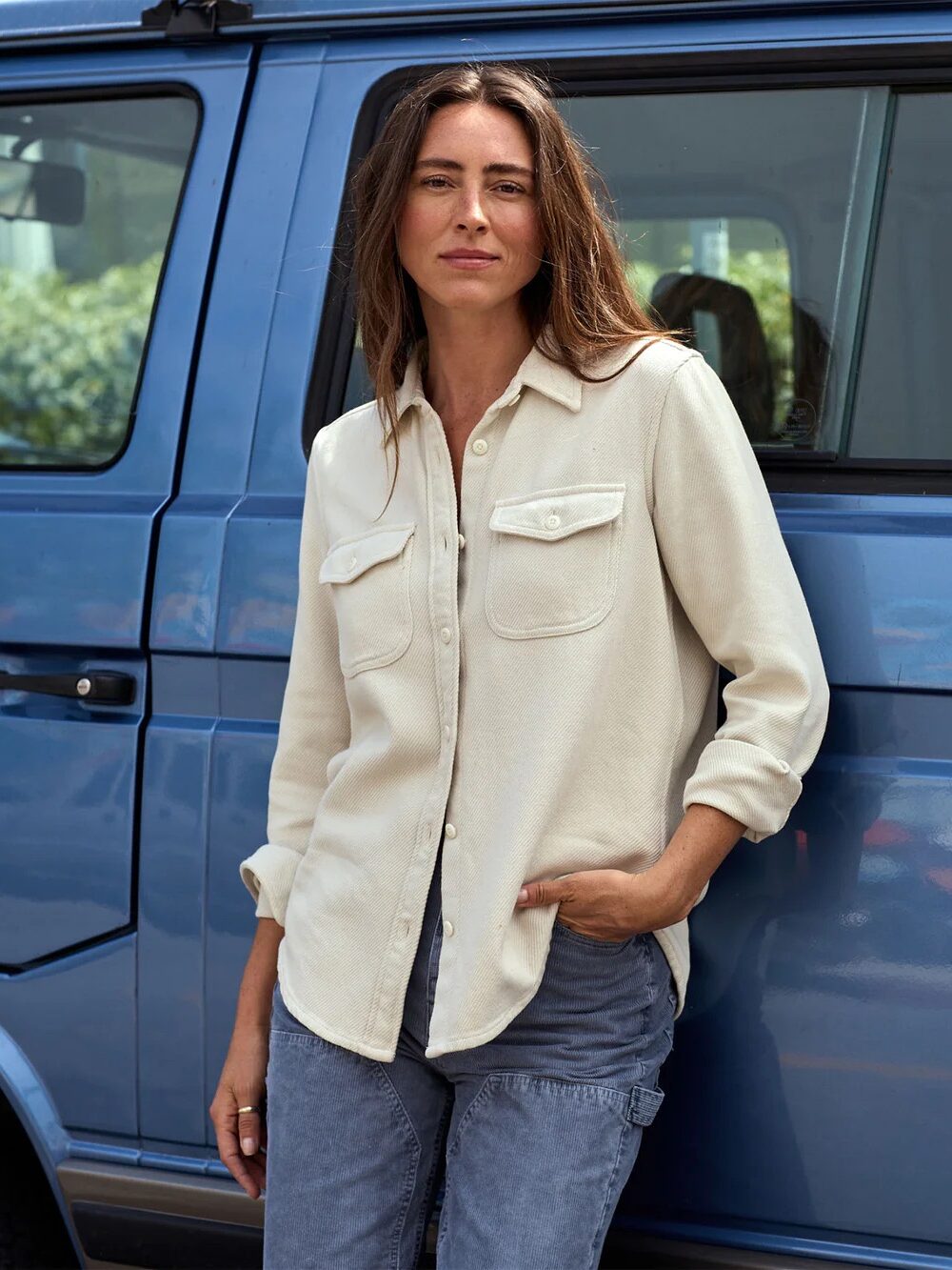 A woman with long brown hair wearing a cream button-up shirt and gray pants stands in front of a blue vehicle, looking at the camera.