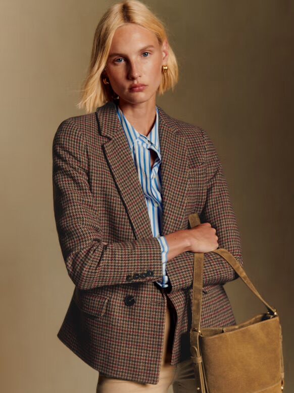 A person with short blonde hair wearing a plaid blazer, blue-striped shirt, and beige pants, holding a tan suede bag against a plain background.