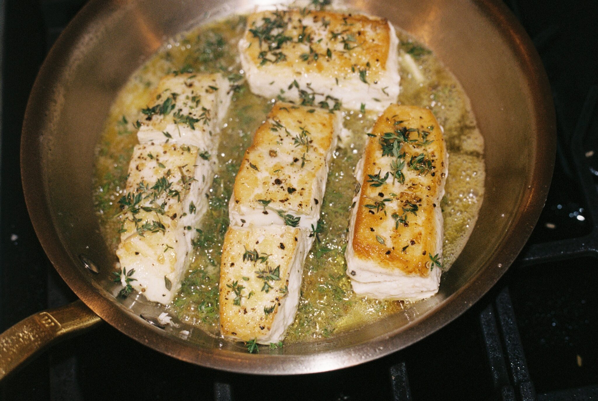 Four pieces of fish are searing in a pan with melted butter and fresh herbs, likely thyme, on a stovetop.