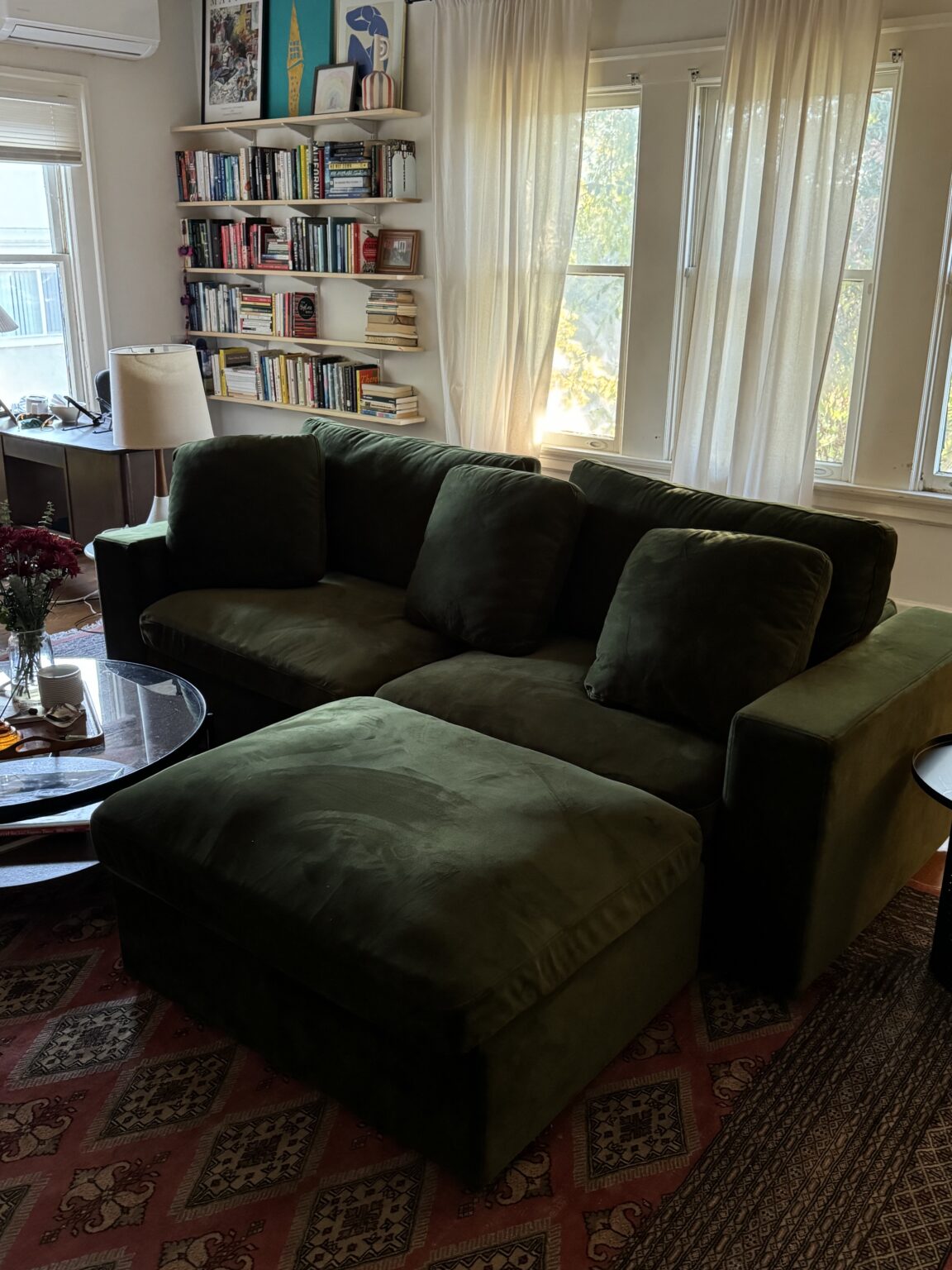 A green velvet sectional sofa sits in a living room with a red patterned rug, bookshelves, a round glass coffee table, and large windows with sheer white curtains.