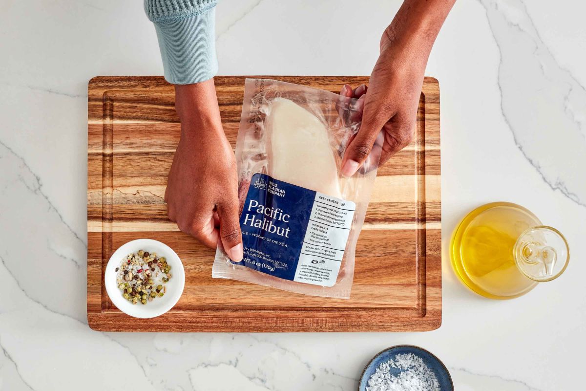 A person places a package of Pacific Halibut on a wooden board. Nearby are a bowl of herbs, a bowl of salt, and a bottle of olive oil.