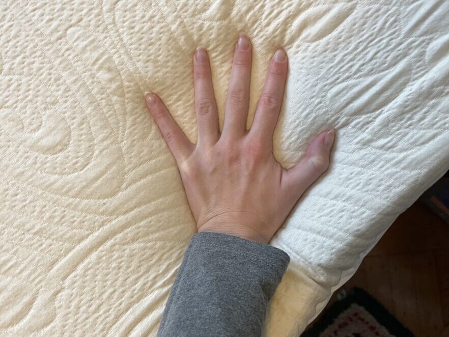 A hand pressing down on the surface of a textured, white organic mattress with a patterned rug partially visible on the floor nearby.