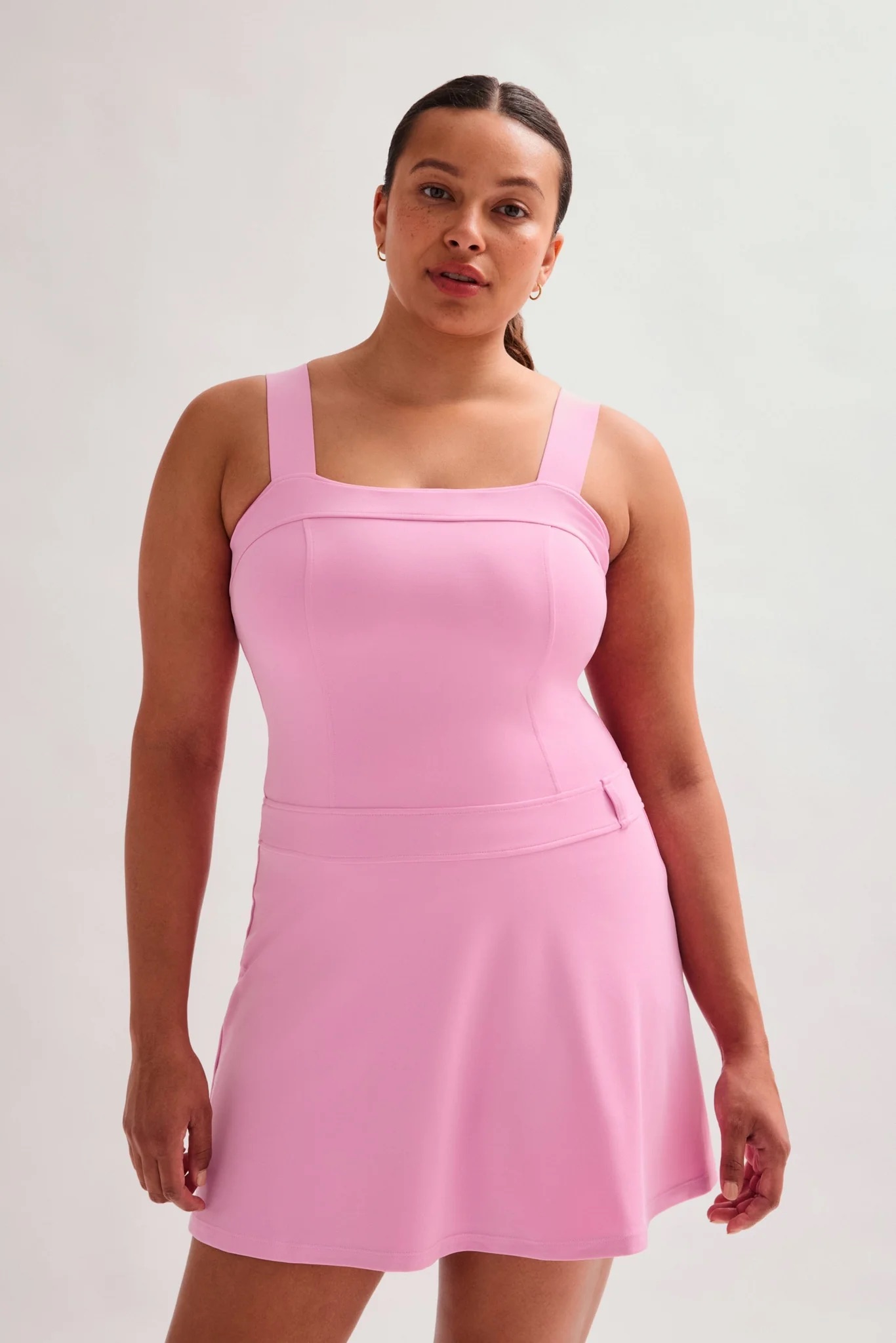 A woman wearing a sleeveless, square-neck, light pink dress stands against a plain light background.