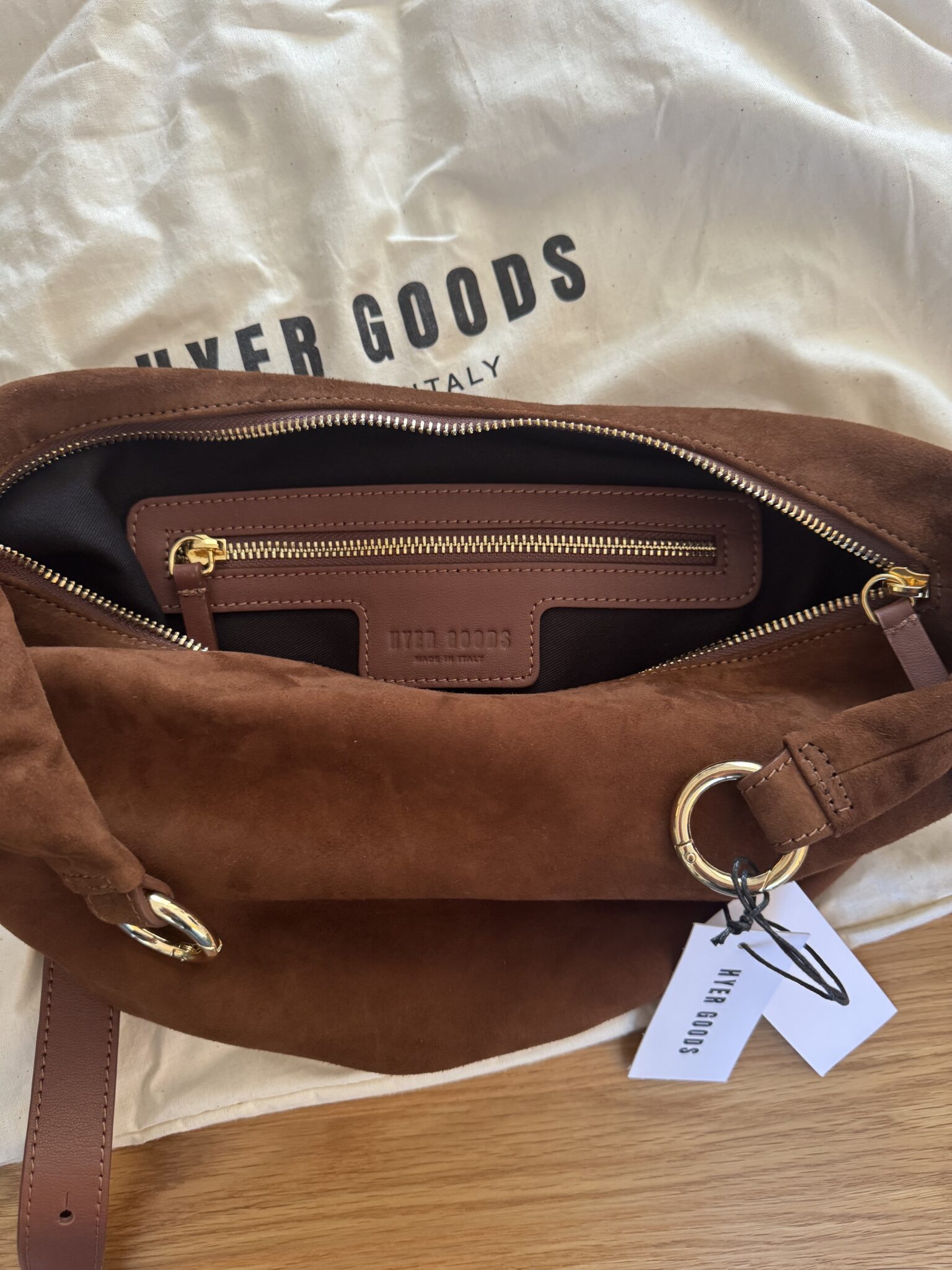A brown suede bag with gold hardware sits open, showing a zippered inner pocket. A white dust bag with "RYER GOODS" printed on it lies underneath.