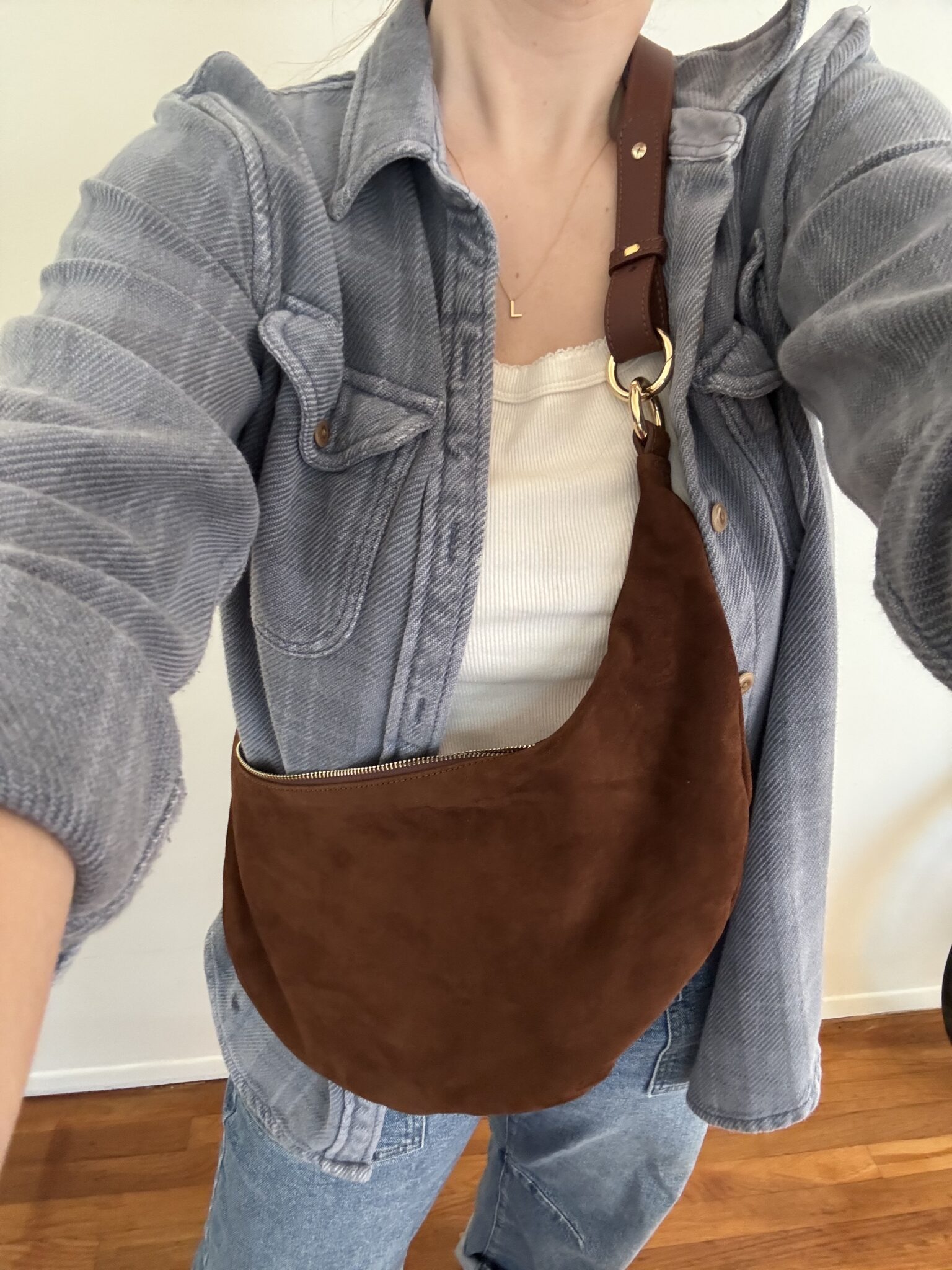 Person wearing a light gray button-up shirt, white top, and blue jeans, carrying a brown suede shoulder bag with a wide strap.
