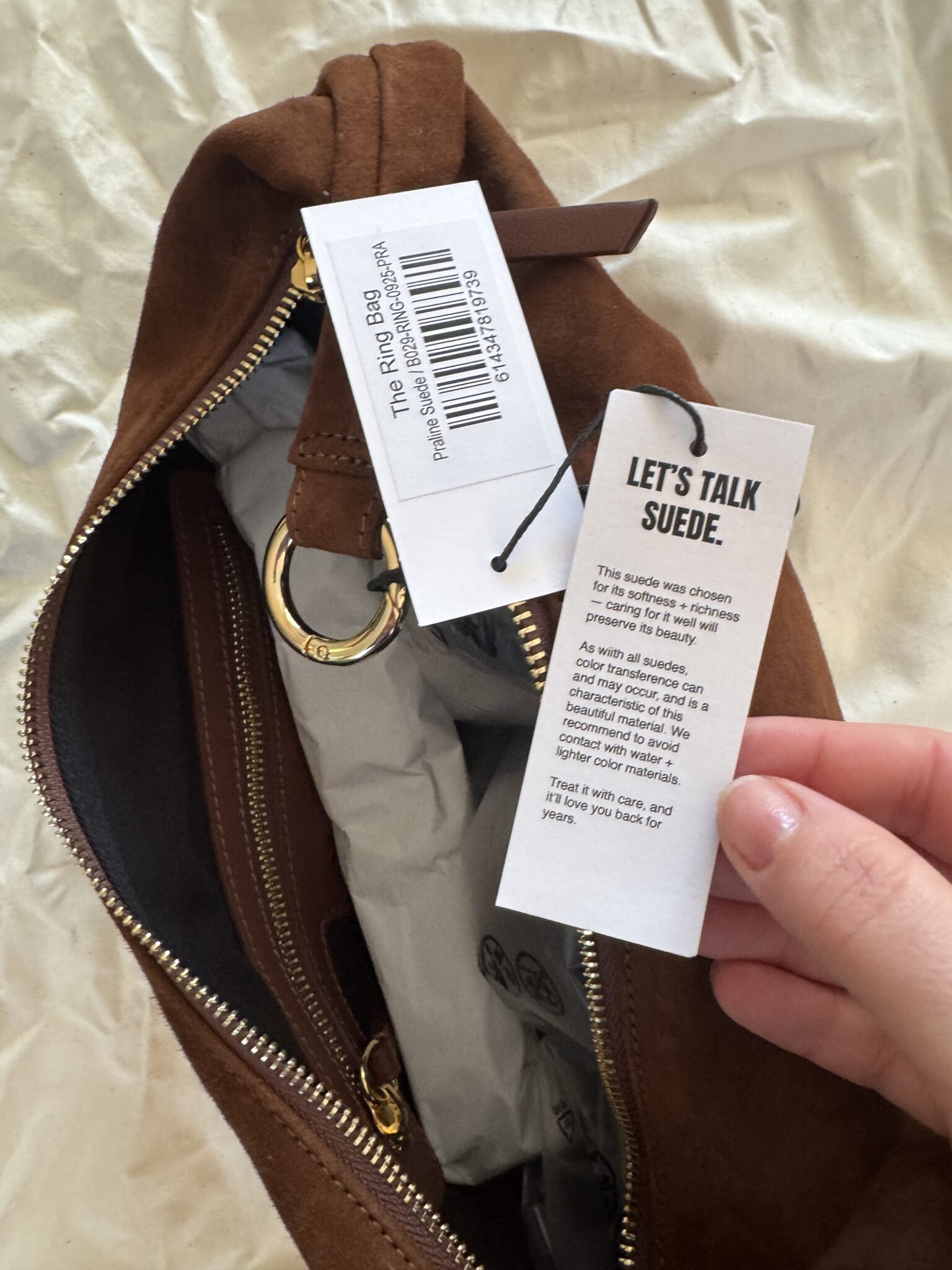 A hand holds a tag inside a brown suede bag. The tag describes the qualities and care of the suede material. A barcode tag labeled "The Ring Bag" is also visible.