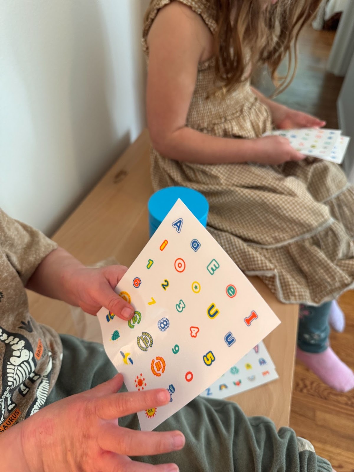 Two children sit on a wooden bench, holding sheets of colorful letter and number stickers. One wears a patterned top and green pants; the other wears a brown checkered dress.