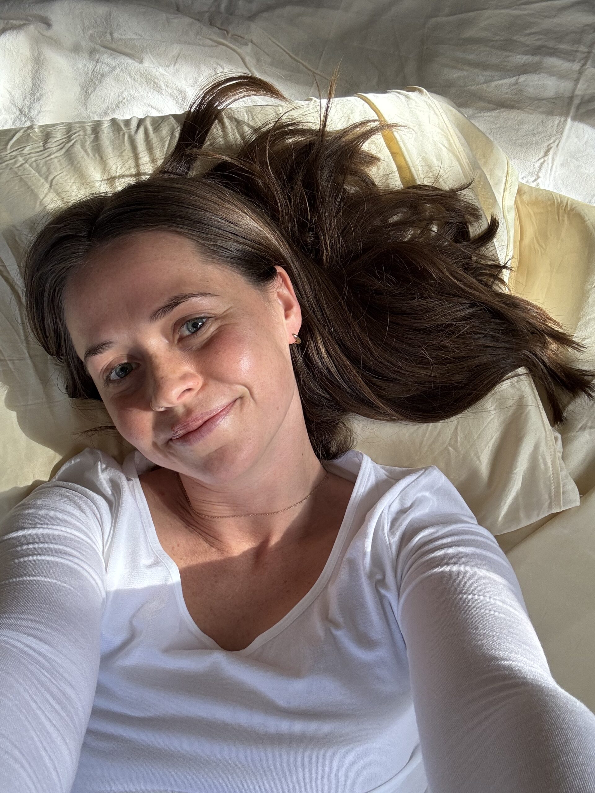 A person with long brown hair wearing a white shirt lies on a bed of cream-colored pillows, smiling at the camera.