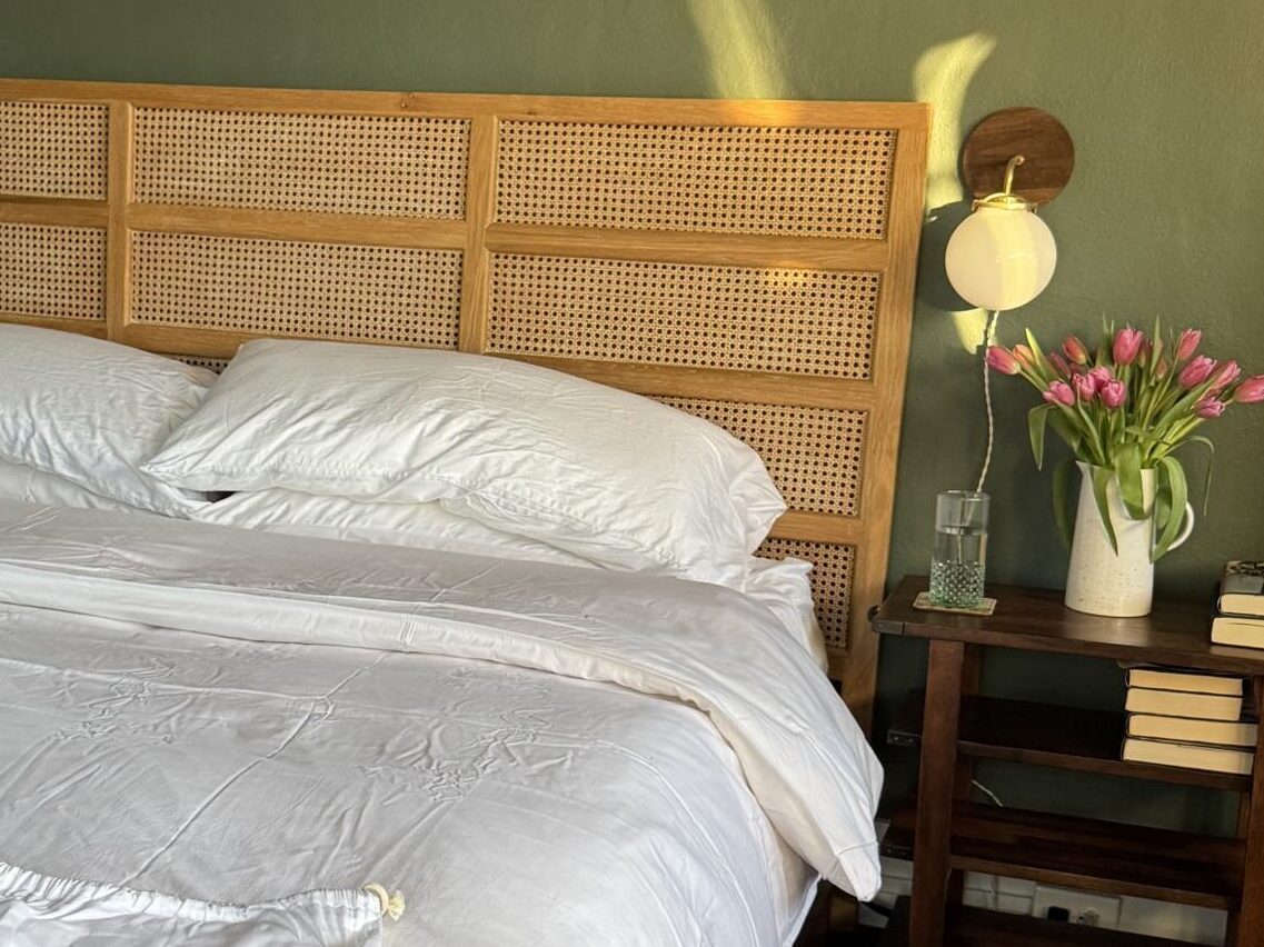A neatly made bed with white linens and a rattan headboard, featuring an organic mattress, a drawstring bag labeled "birch," and a bedside table with books, flowers, and a glass of water.