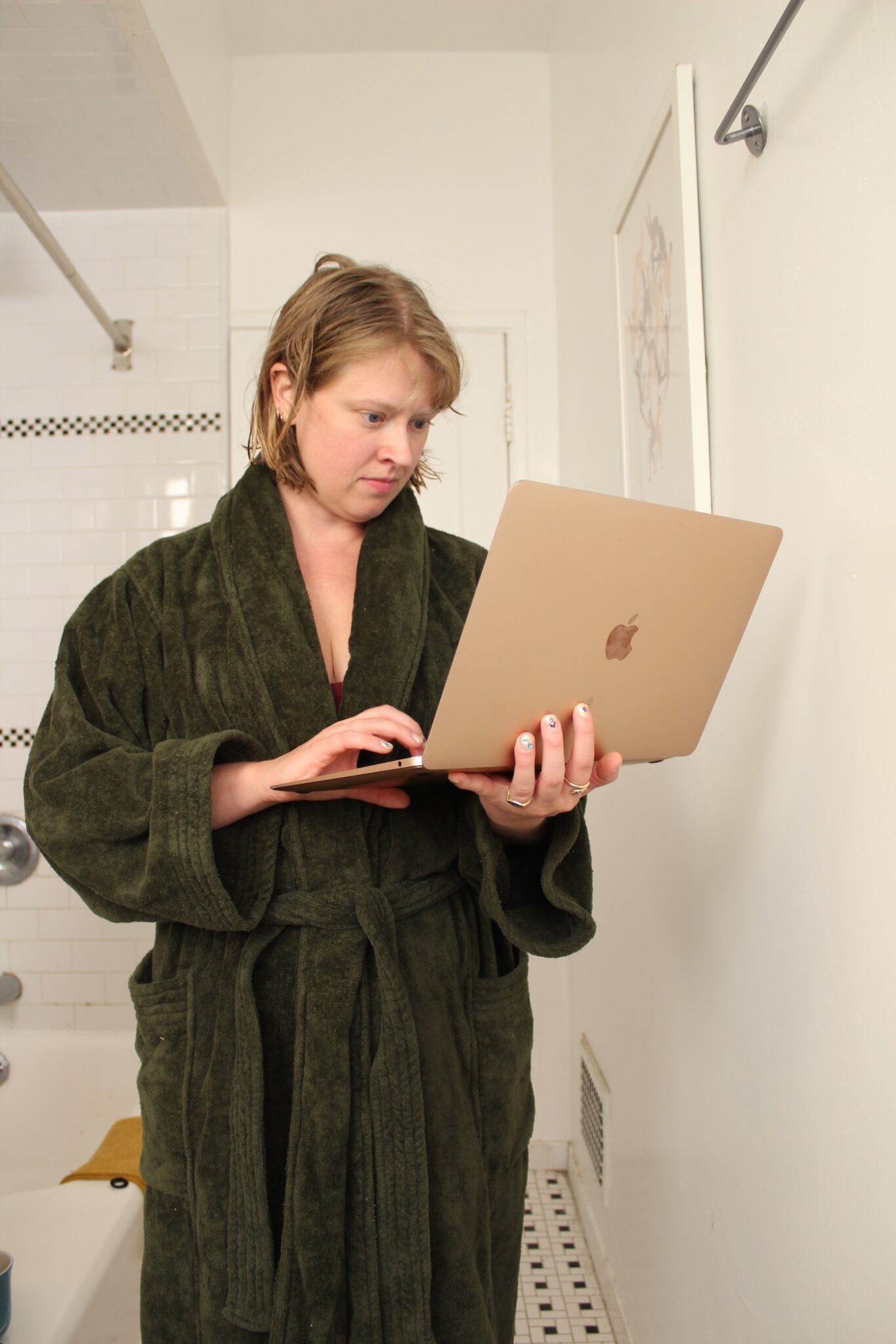 A person in a dark green bathrobe stands in a bathroom, looking at and typing on a laptop.