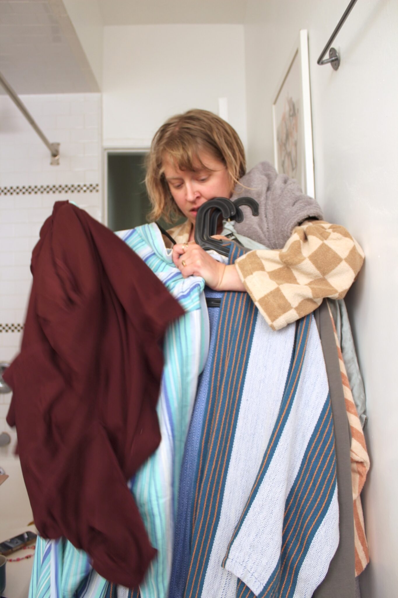 A woman stands in a bathroom, holding several clothes and towels on hangers, appearing to be sorting or carrying laundry.