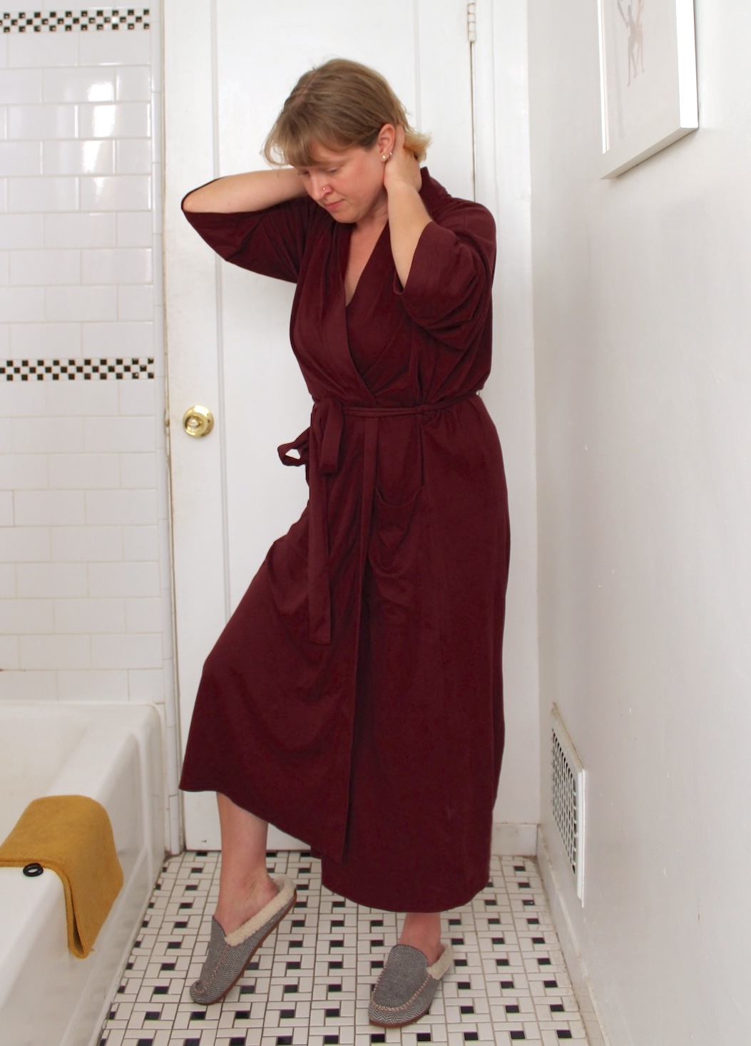 Person in a maroon robe and gray slippers stands in a white tiled bathroom, touching their neck with both hands near a closed door.
