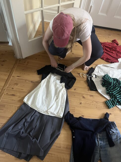 A person in a red cap arranges clothes on a wooden floor, laying out tops, skirts, and sweaters in coordinated outfits near a doorway.