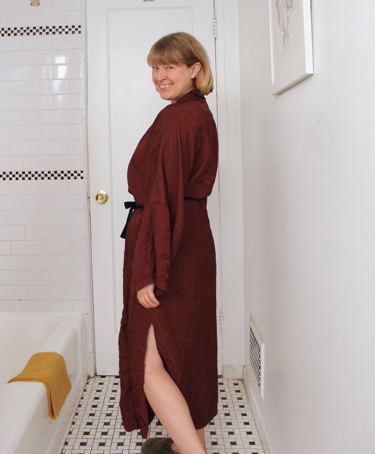 Person standing sideways in a bathroom, wearing a long maroon robe with a slit; white tiled walls and floor, a bathtub, and a towel are visible.